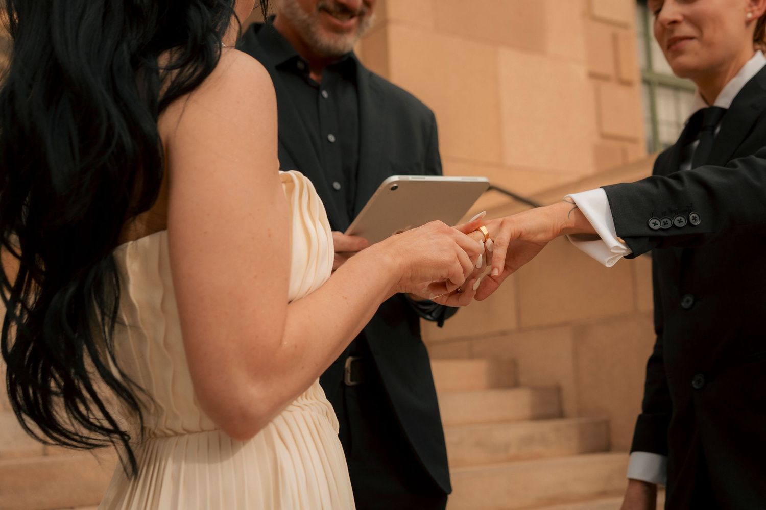 Bride and groom exchange rings during intimate outdoor wedding ceremony with officiant holding tablet.