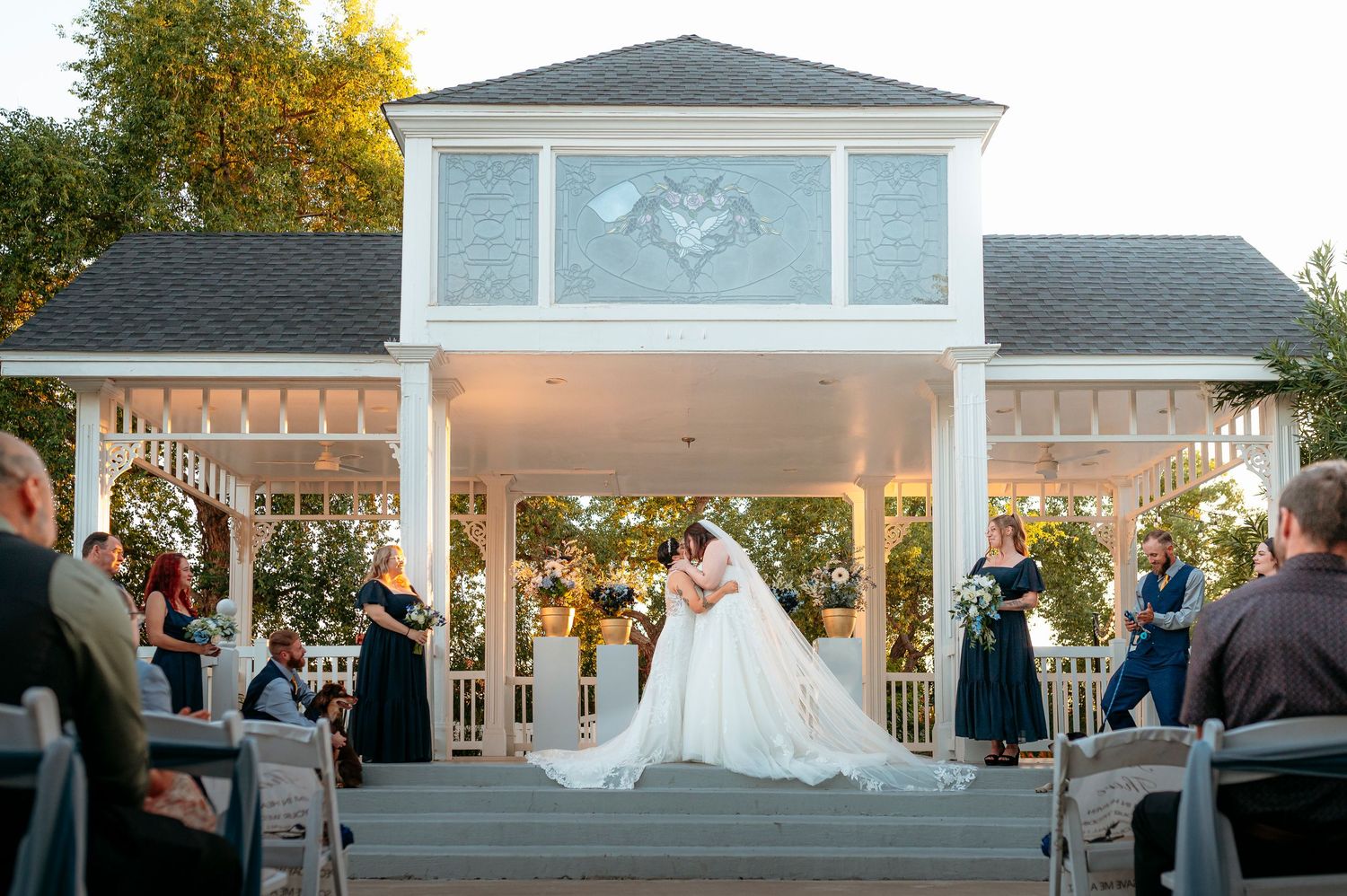 Bride and groom kiss at the altar of a white gazebo ceremony venue surrounded by seated wedding guests.