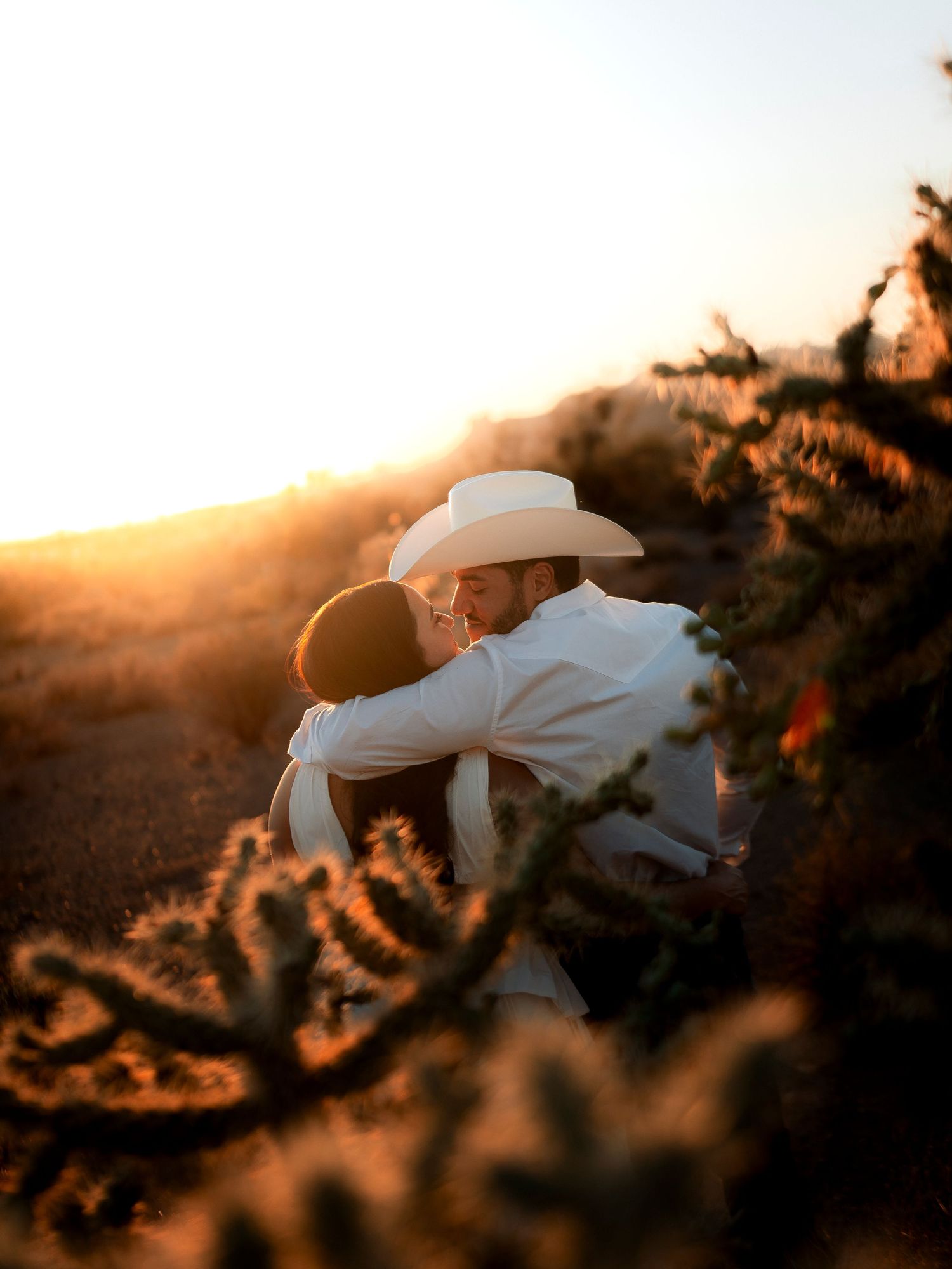 Couple embracing at golden hour in a desert landscape with cacti, bathed in warm sunset light.
