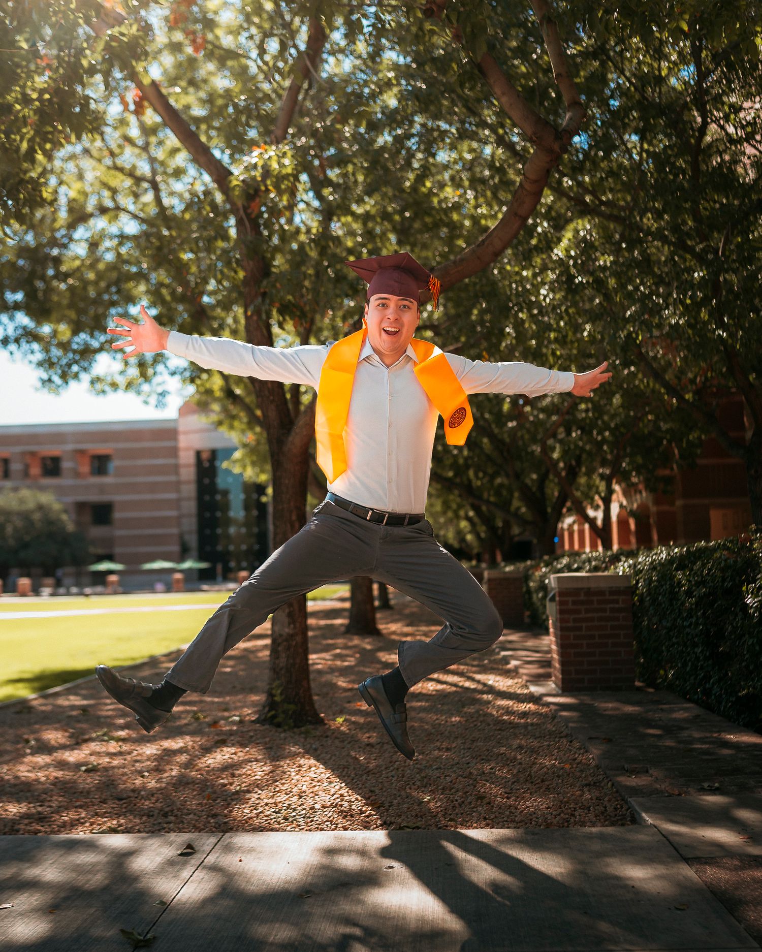 Jubilant graduate jumps with arms spread wide wearing maroon cap and gold stole outdoors.