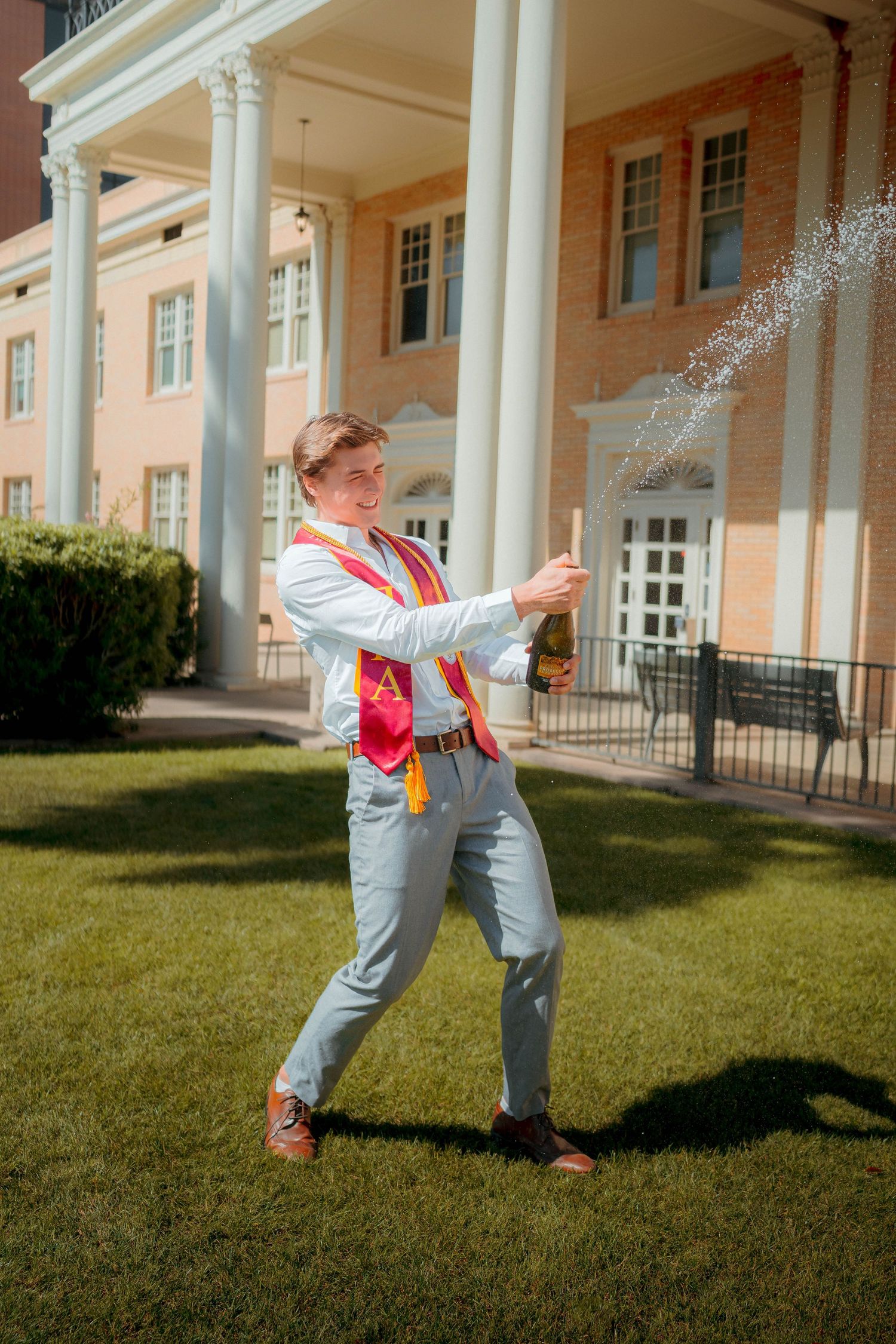 Graduate in light blue suit sprays champagne bottle outdoors near white-columned brick building on sunny day.