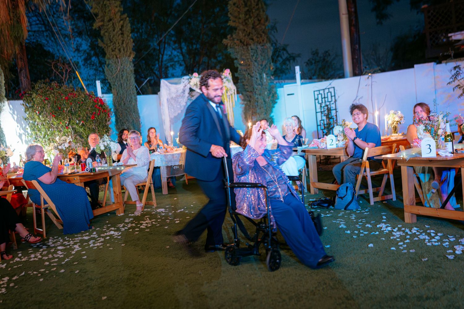 Groom pushing elderly woman in wheelchair at outdoor evening wedding reception.