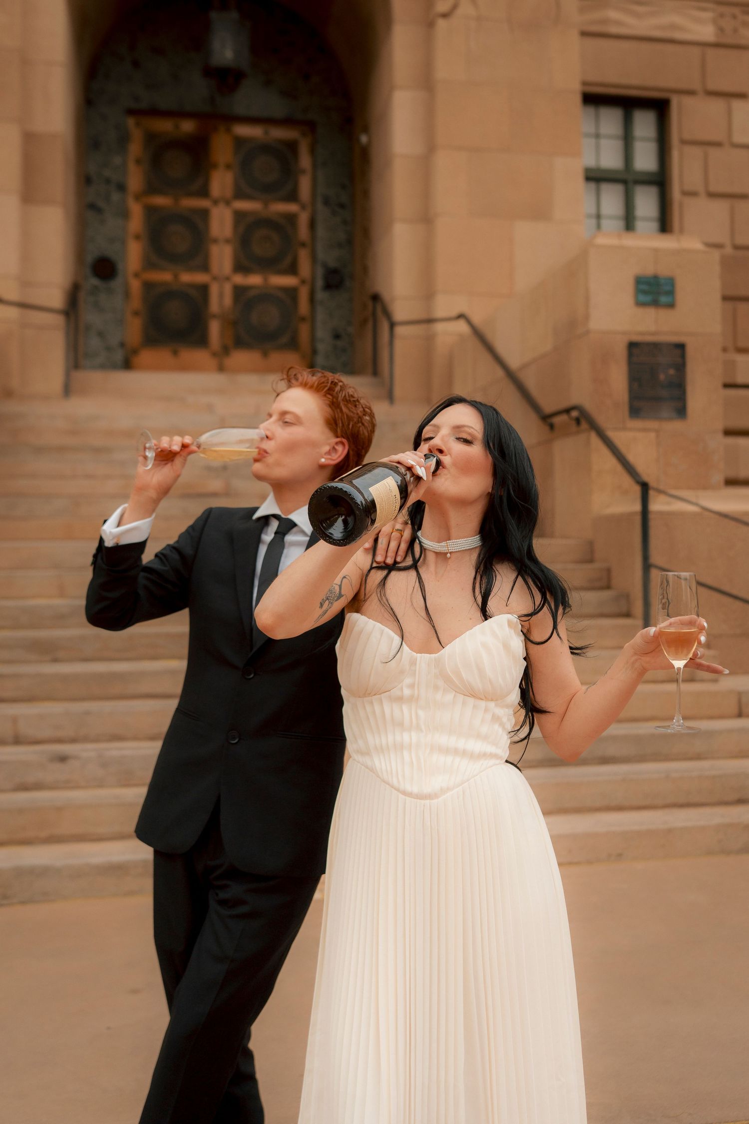 Bride and groom drinking champagne from bottle outside historic building on wedding day.