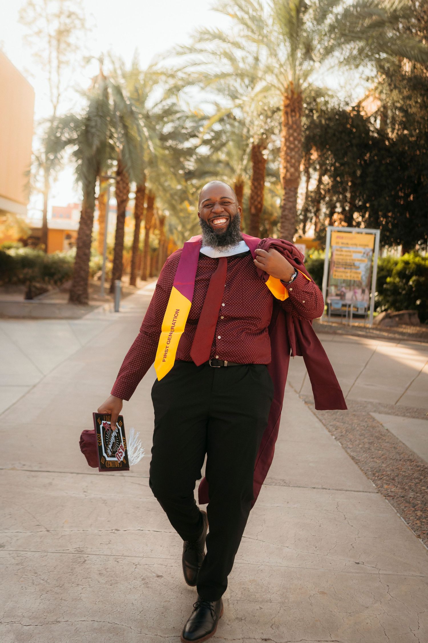 A joyful graduate in maroon gown walks among palm trees holding his diploma.