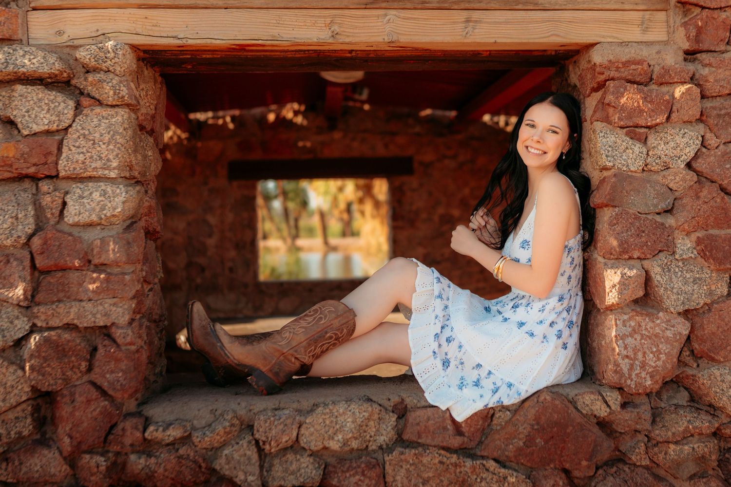 Smiling young woman in floral dress and cowboy boots sitting in a rustic stone alcove.