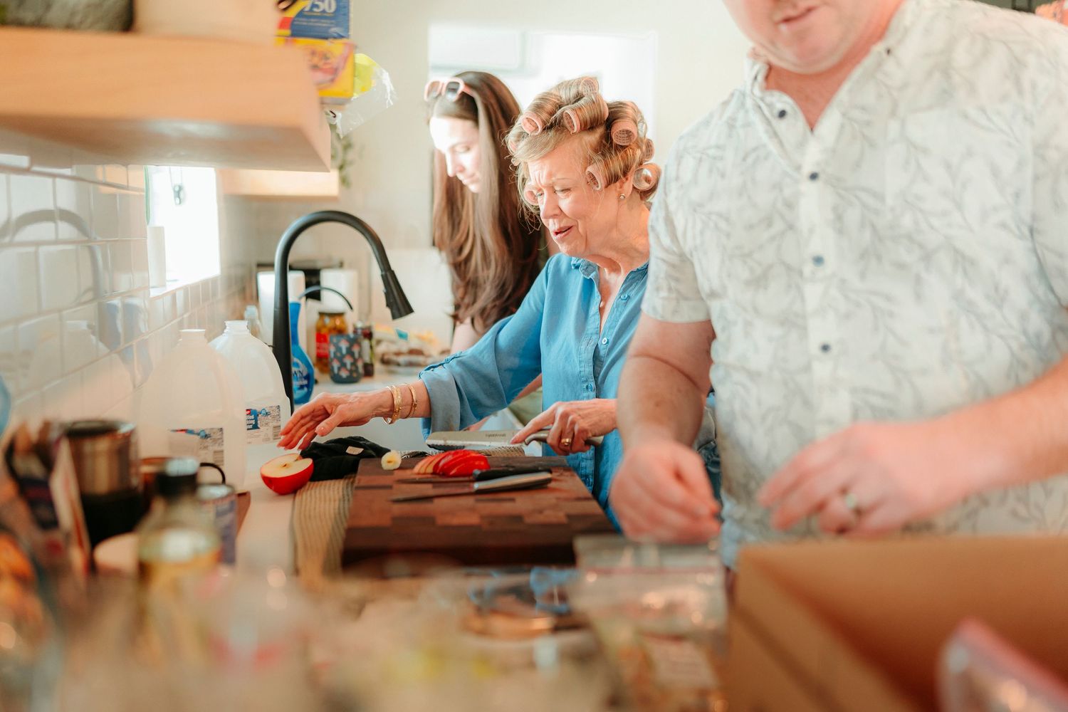 Elderly woman with hair curlers chopping vegetables on wooden cutting board in busy kitchen with family.