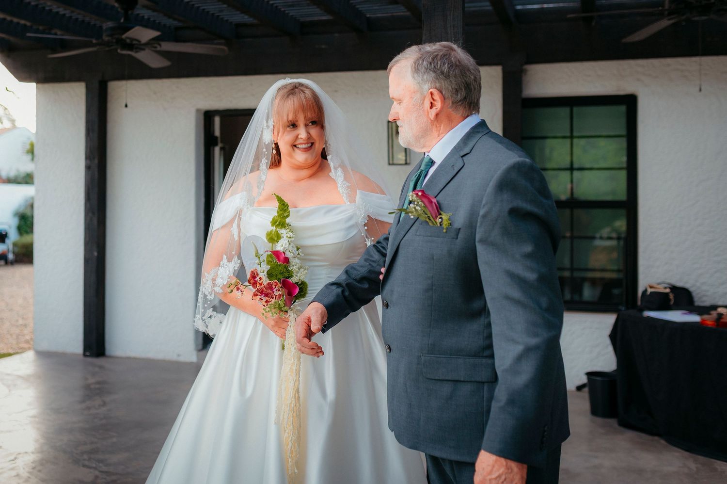 Bride in white gown and veil holds red bouquet while sharing a tender moment with a man in a gray suit at a wedding.