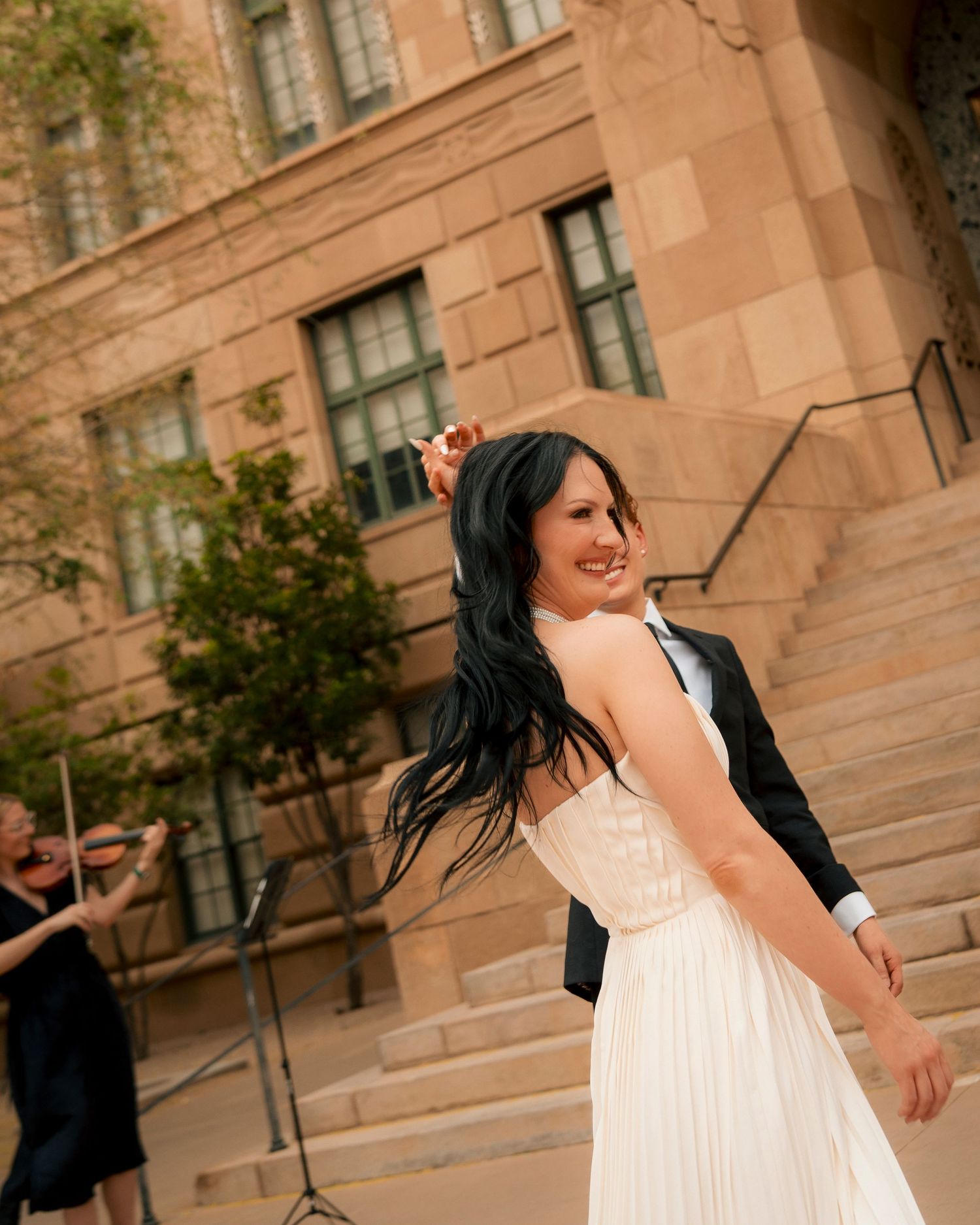 Bride in white gown twirls with groom outside stone building as violinist performs in background.