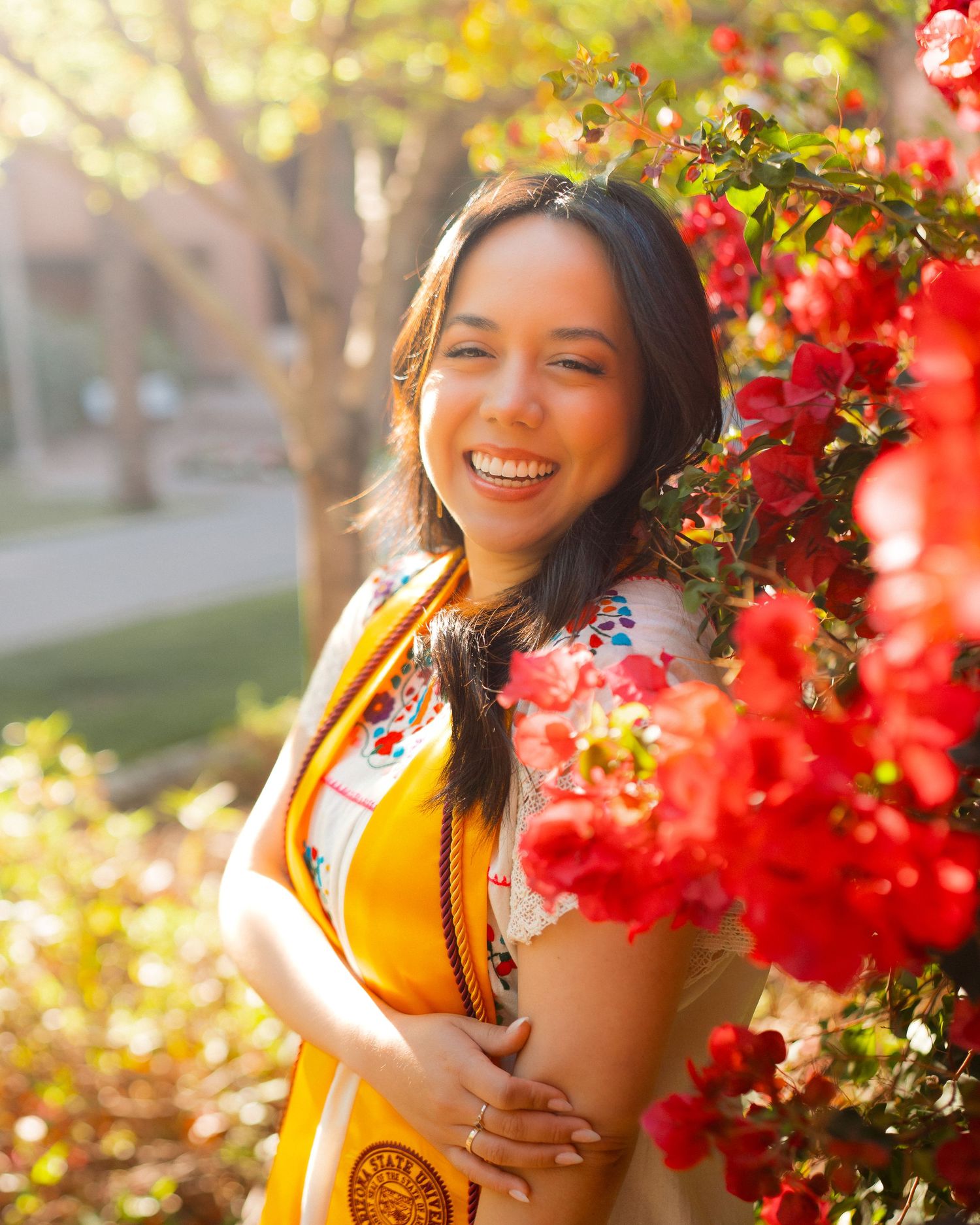 Smiling young woman in a yellow graduation sash posing beside vibrant red bougainvillea flowers.
