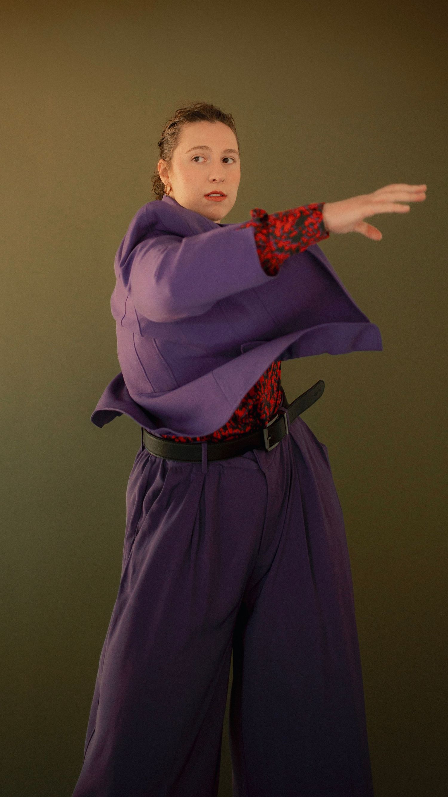 Person posing in purple wide-leg pants and jacket with red floral top and black belt against green background.