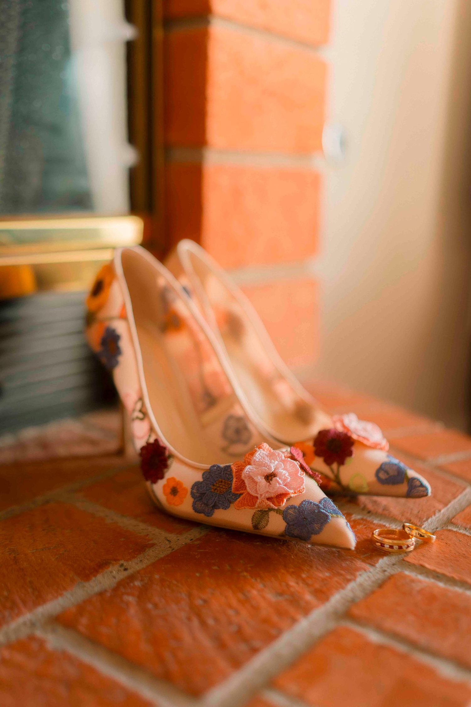 Elegant floral embroidered wedding heels with pink and blue flowers displayed on rustic brick surface.