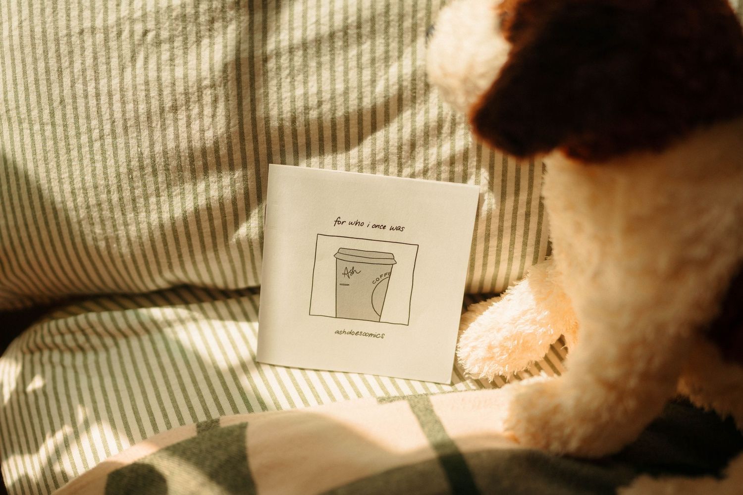 Ash Does Comics' illustrated zine resting against a fluffy teddy bear on striped bedding.