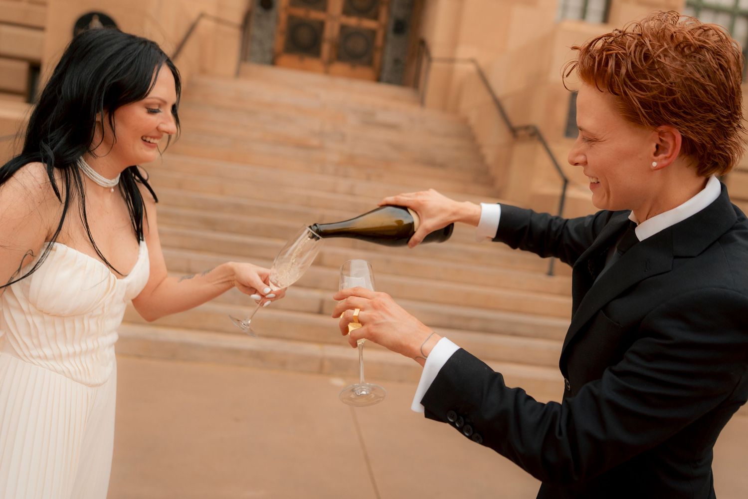 Couple in wedding attire pouring champagne into glasses on stone steps, celebrating their special day.