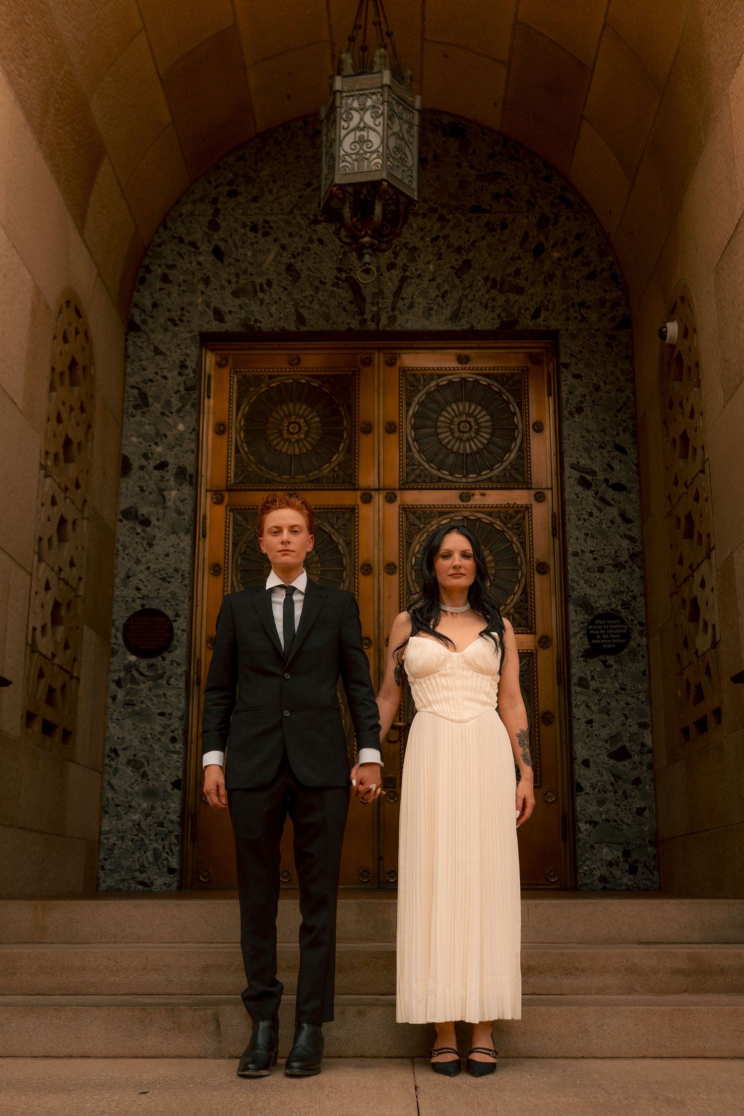 Bride and groom pose solemnly before ornate golden doors of Phoenix City Hall.
