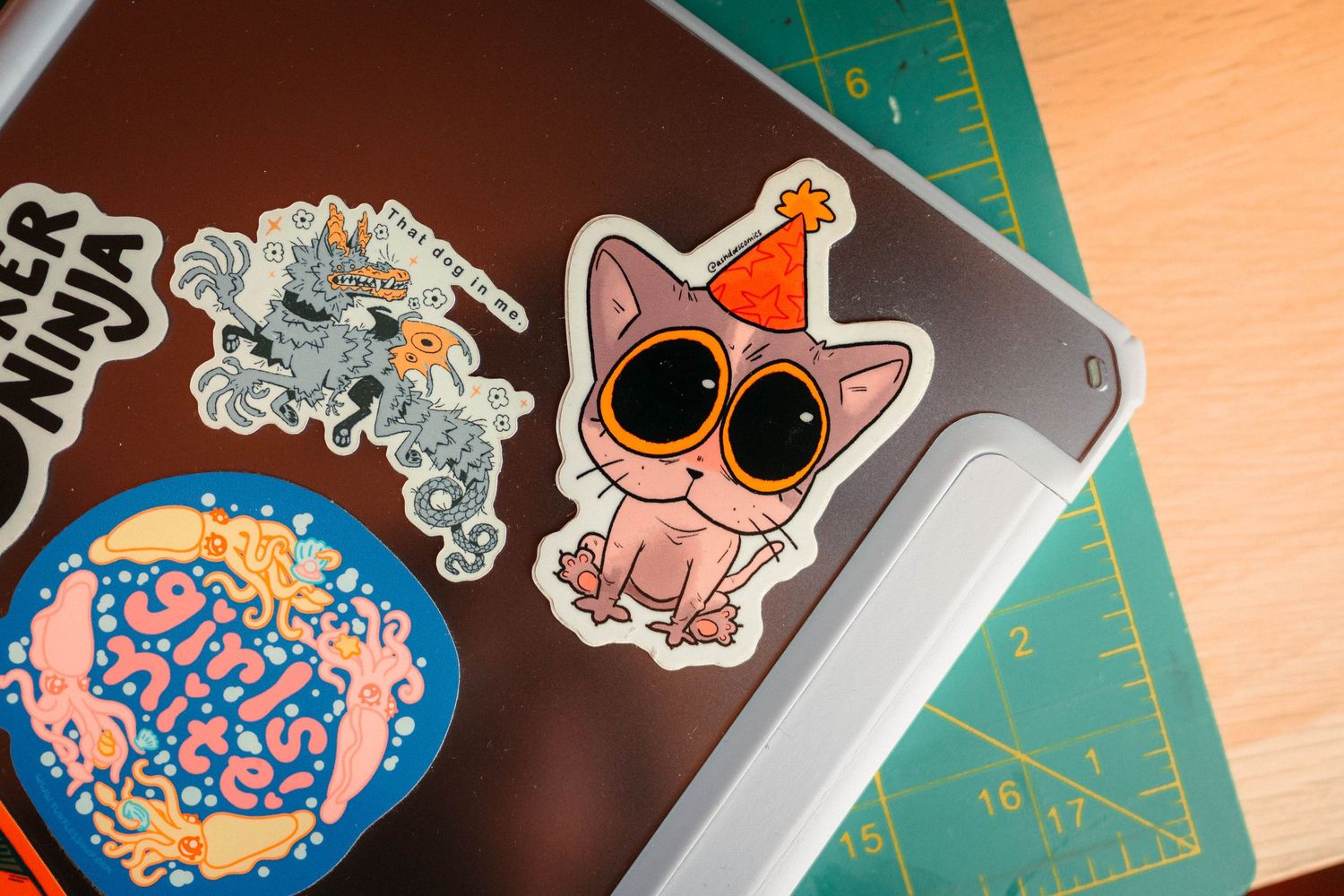 Cute cat with party hat sticker and colorful brain sticker on a cutting mat surface.