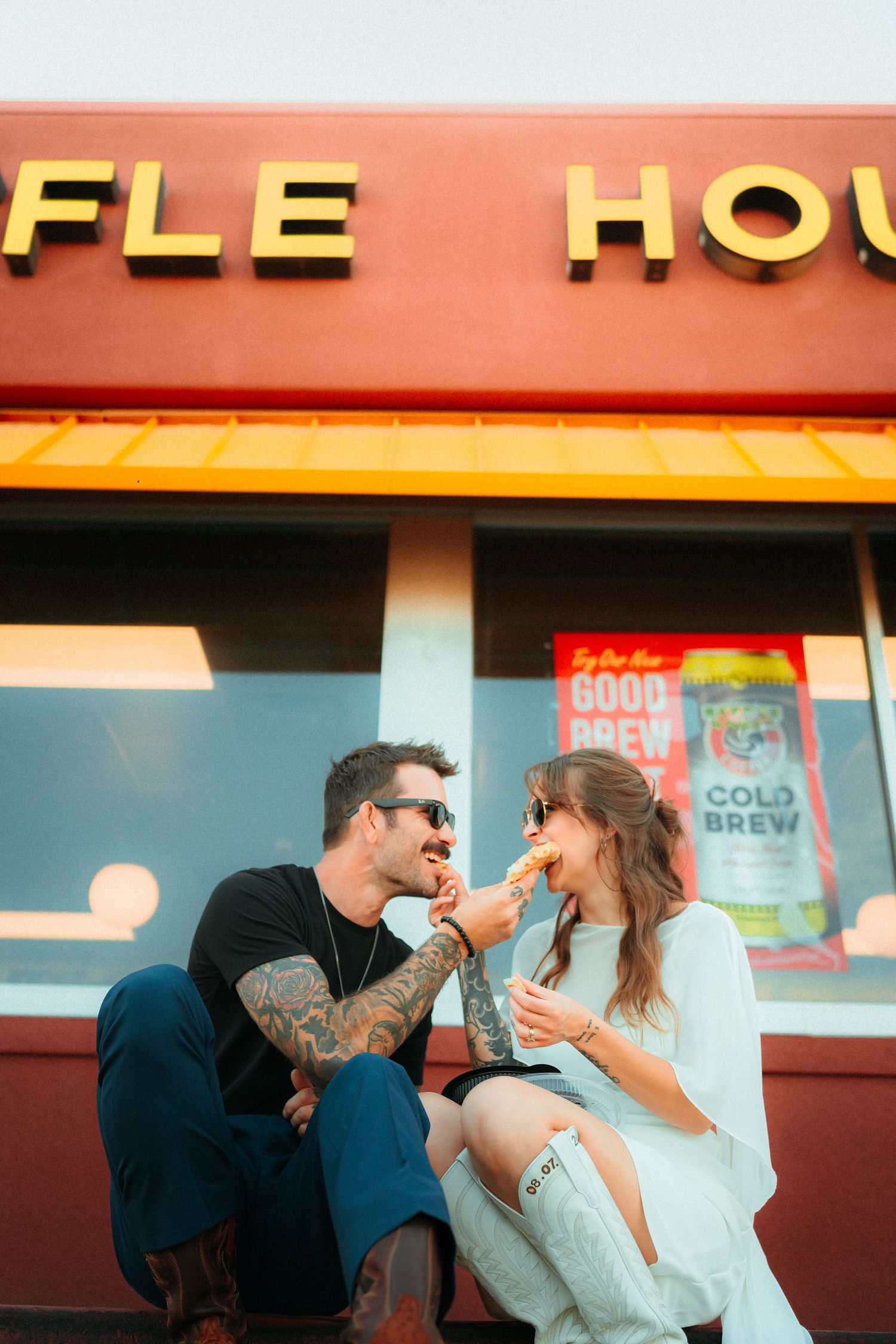 Couple sharing food and laughing outside a Waffle House restaurant, man with tattoos and woman in white dress.
