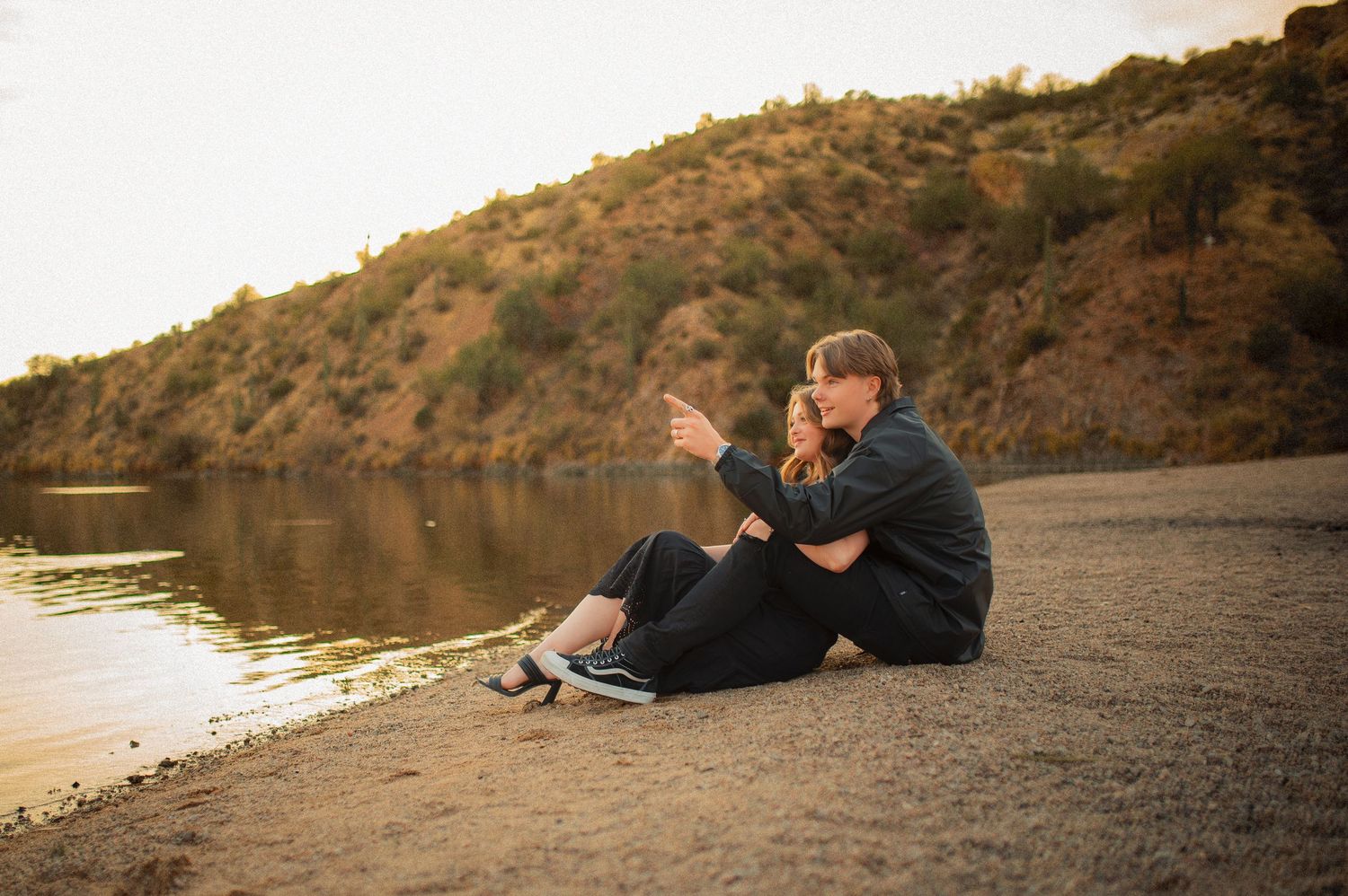 Couple sitting together on lakeside shore at sunset, pointing toward scenic mountain view.