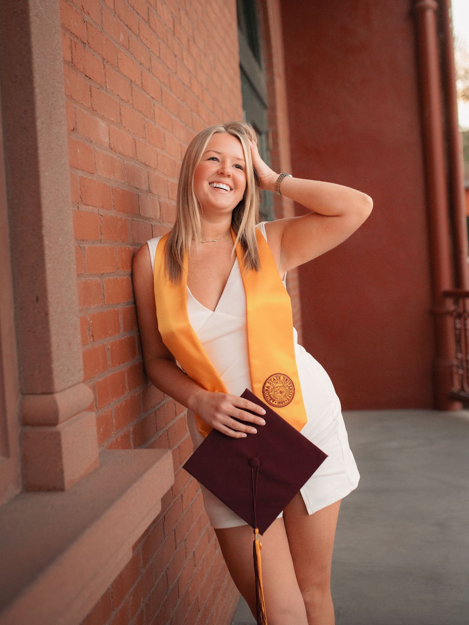 A laughing graduate in white dress leans against brick wall holding maroon cap.