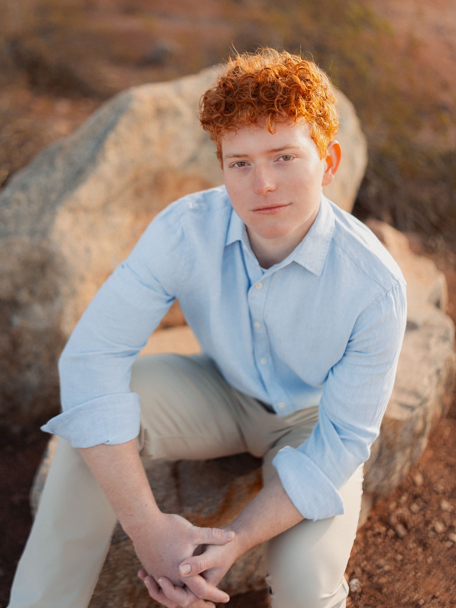 Young man with curly red hair sits on rocks wearing a light blue shirt in a warm outdoor setting.