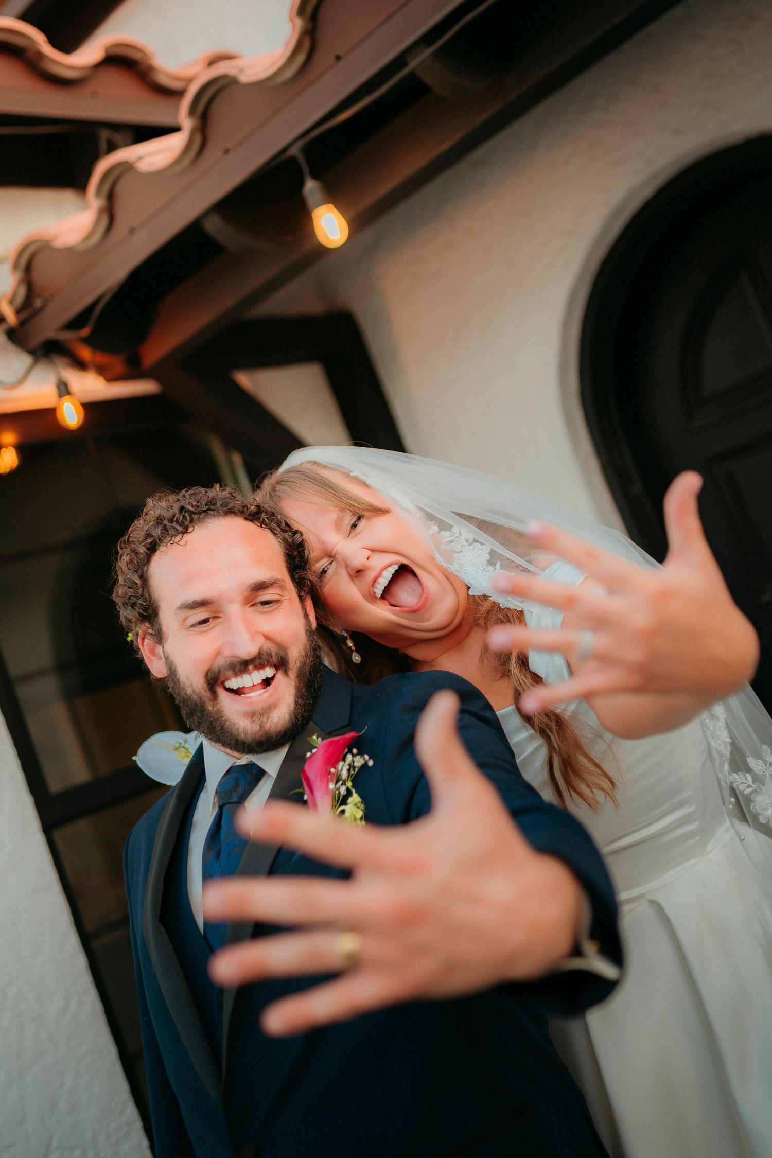 Joyful newlyweds showing off wedding rings, bride in veil laughing, groom in navy suit with boutonniere, at outdoor venue.