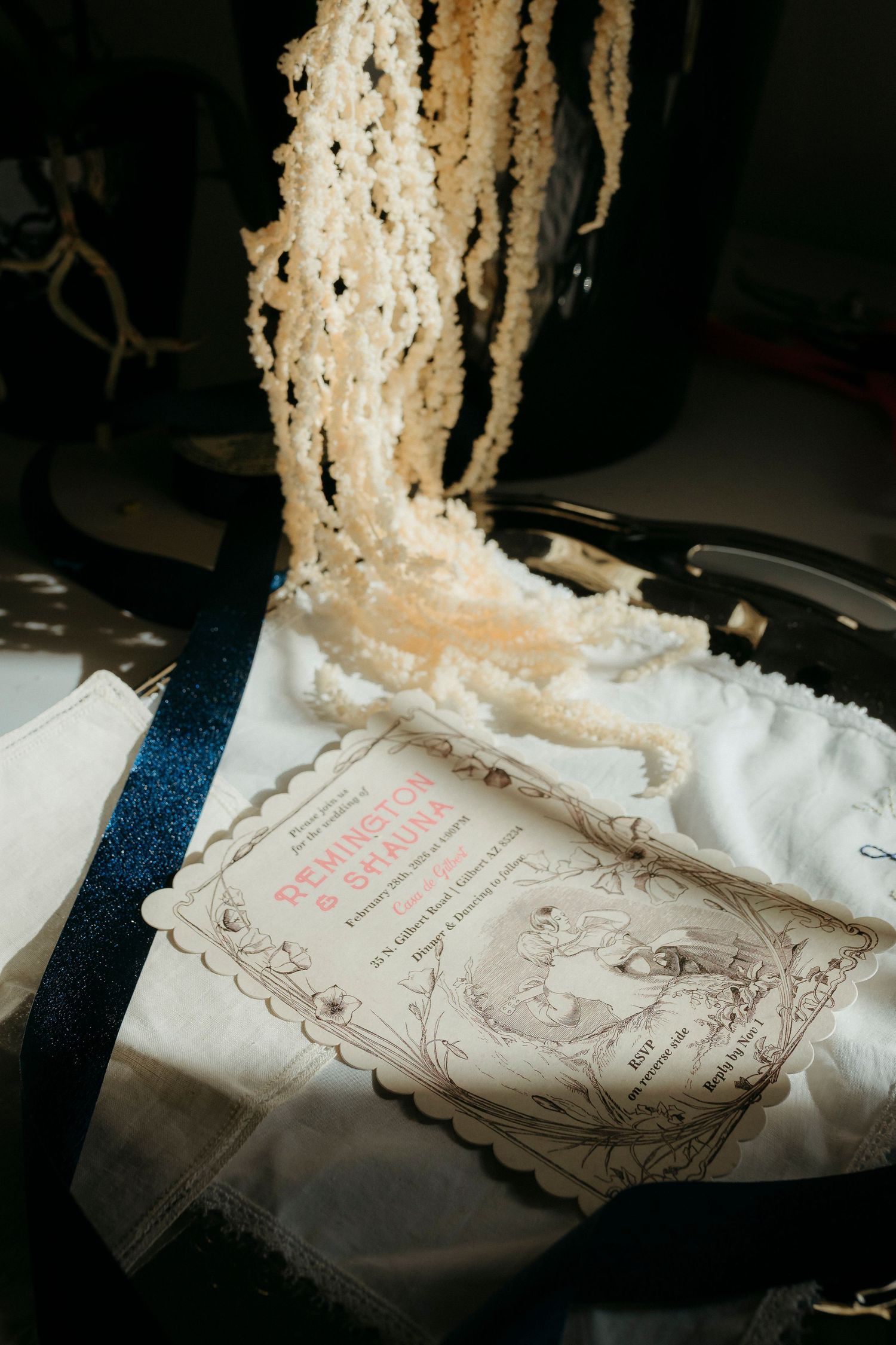 Wedding invitation with pearl bead necklaces and navy ribbon on white fabric in soft natural light.