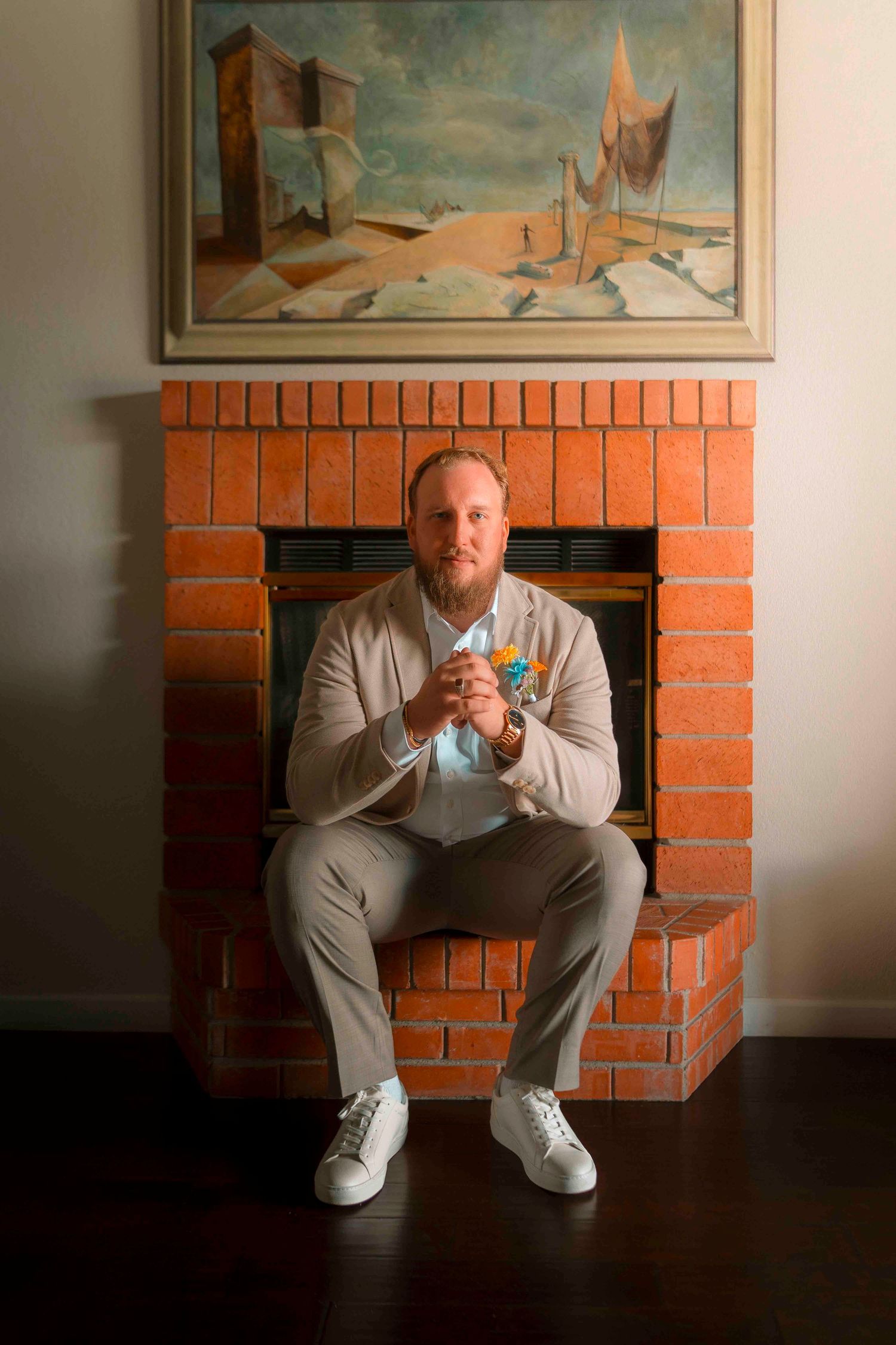Groom in beige suit sitting confidently before a brick fireplace with a desert landscape painting above.