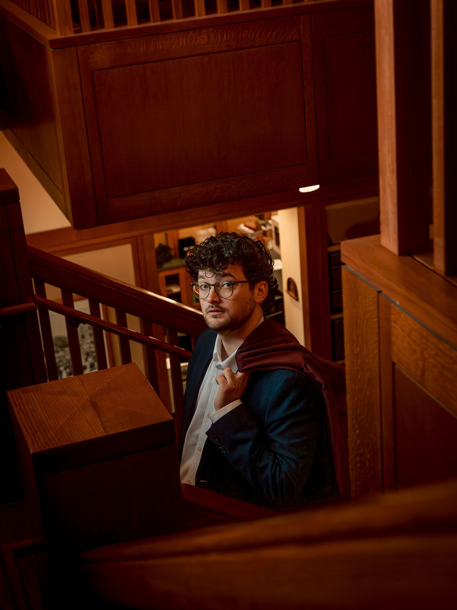 Young man in dark blazer and glasses poses on wooden staircase in warm ambient lighting.