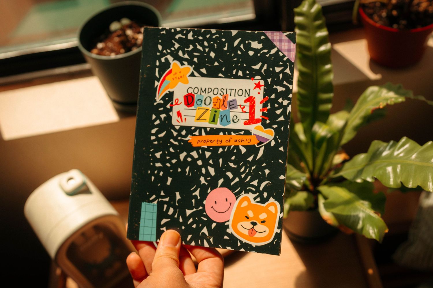Hand holding a dark composition notebook with colorful animal stickers near plants - Ash Does Comics.