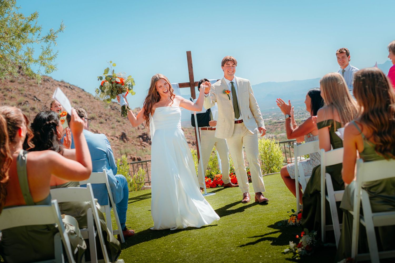 Newlyweds walk hand-in-hand past cheering guests at outdoor desert wedding ceremony under blue sky.