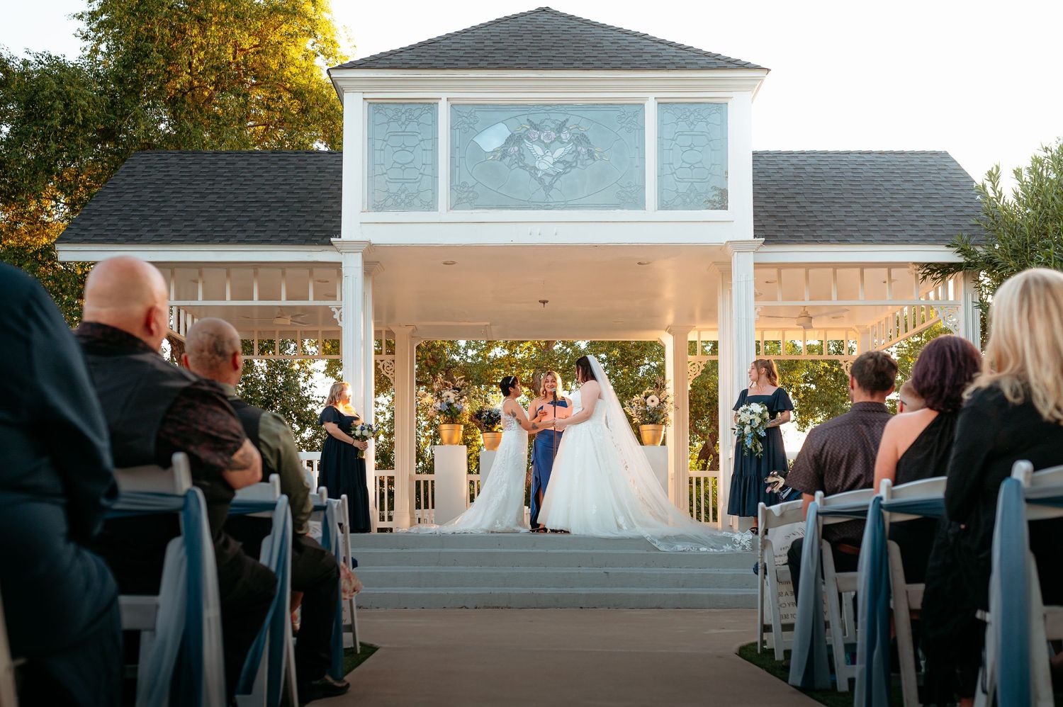 Couple exchanges vows at a white gazebo wedding venue with seated guests in attendance.
