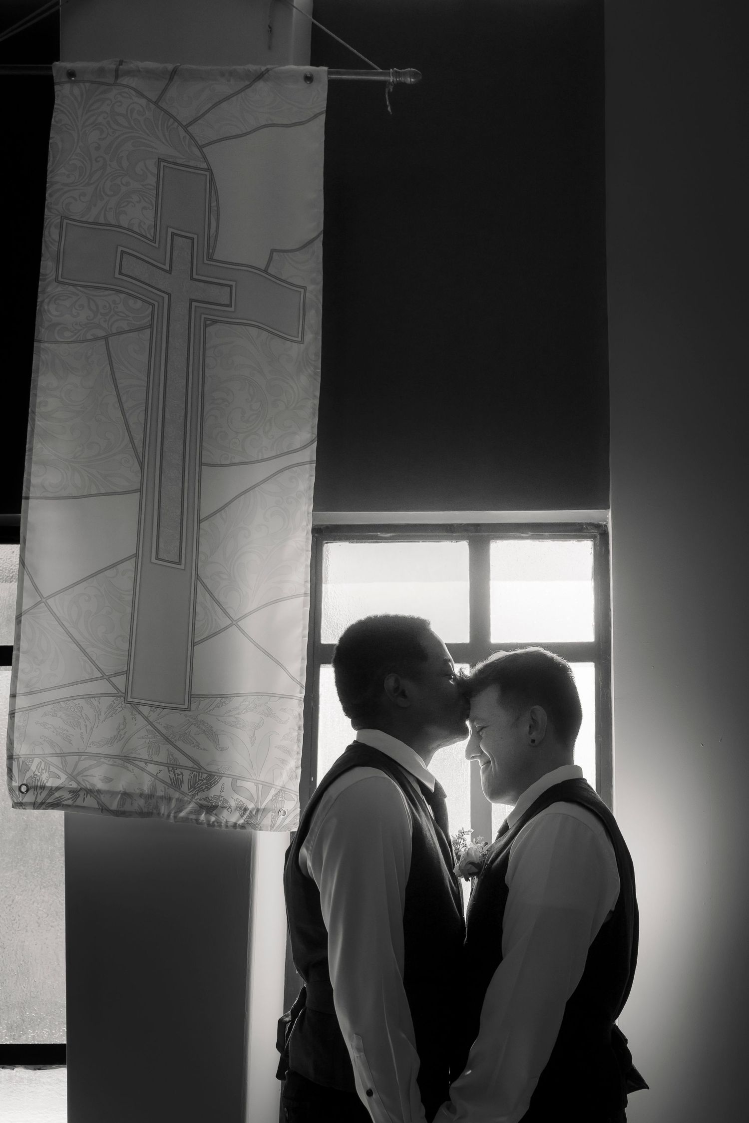 Black and white silhouette of two grooms sharing an intimate moment beneath a cross banner in church.