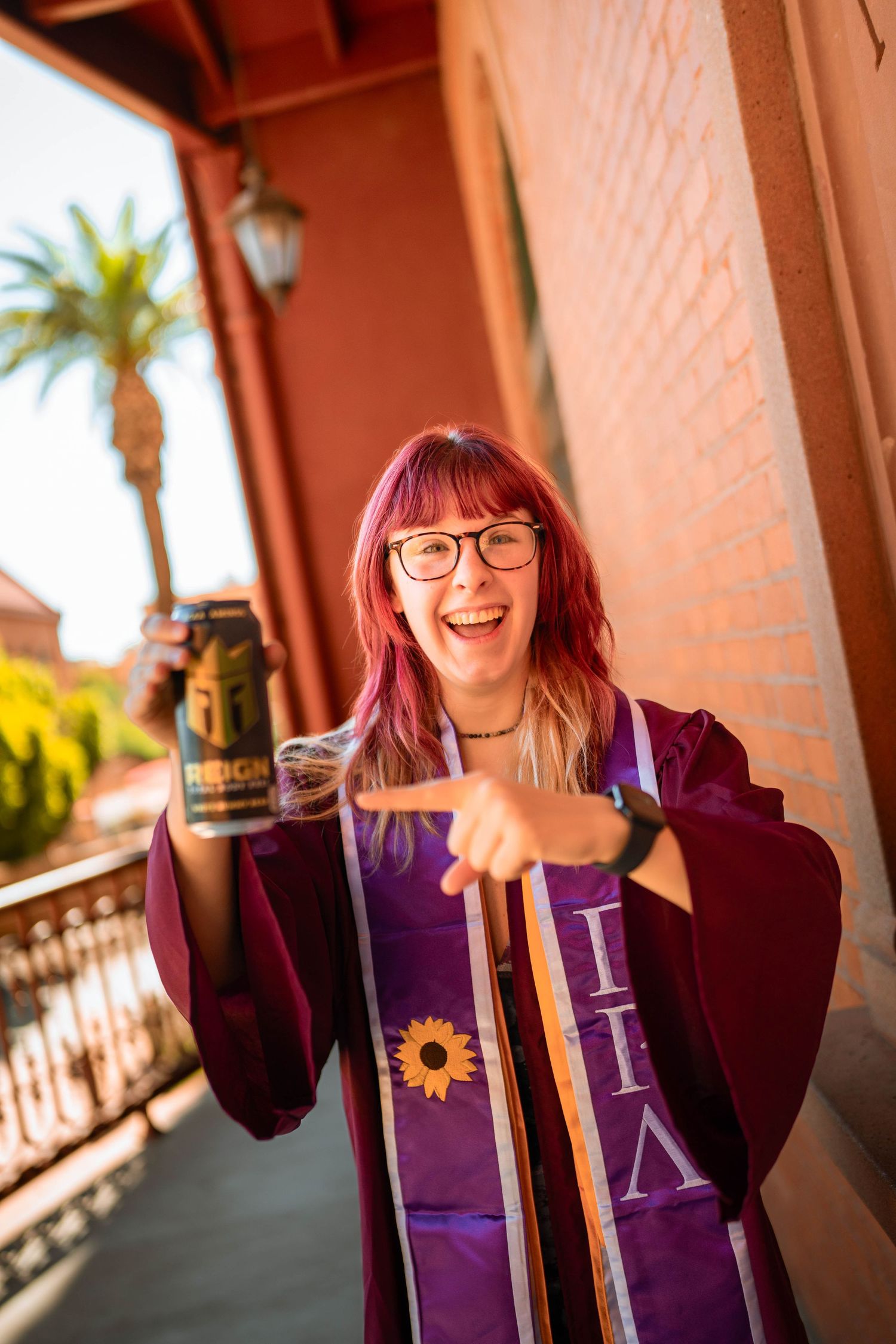 Smiling graduate in maroon cap and gown with purple honor sash holding a mug outdoors.