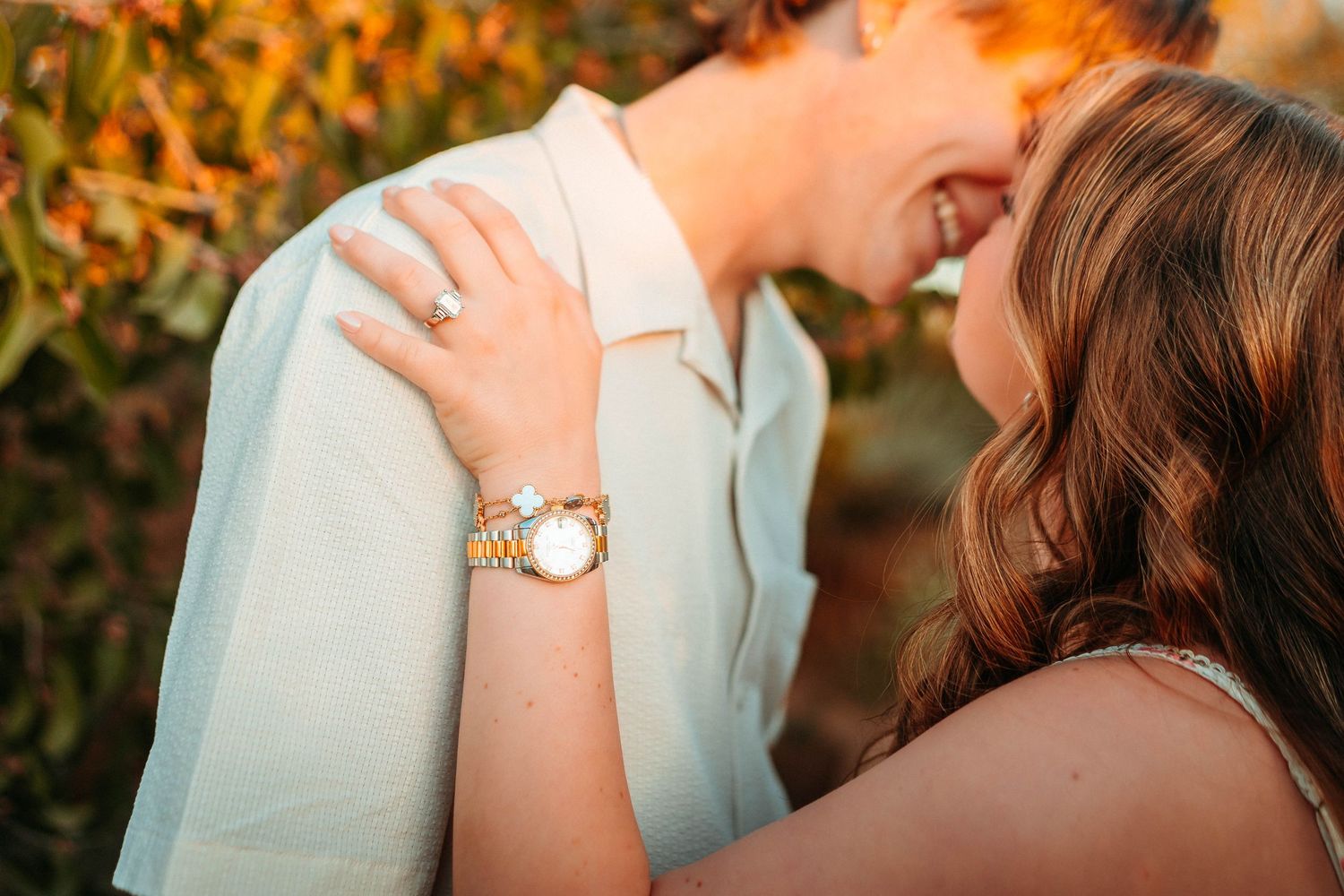 Couple sharing a romantic moment, woman wearing a gold watch and diamond engagement ring resting her hand on man's shoulder.