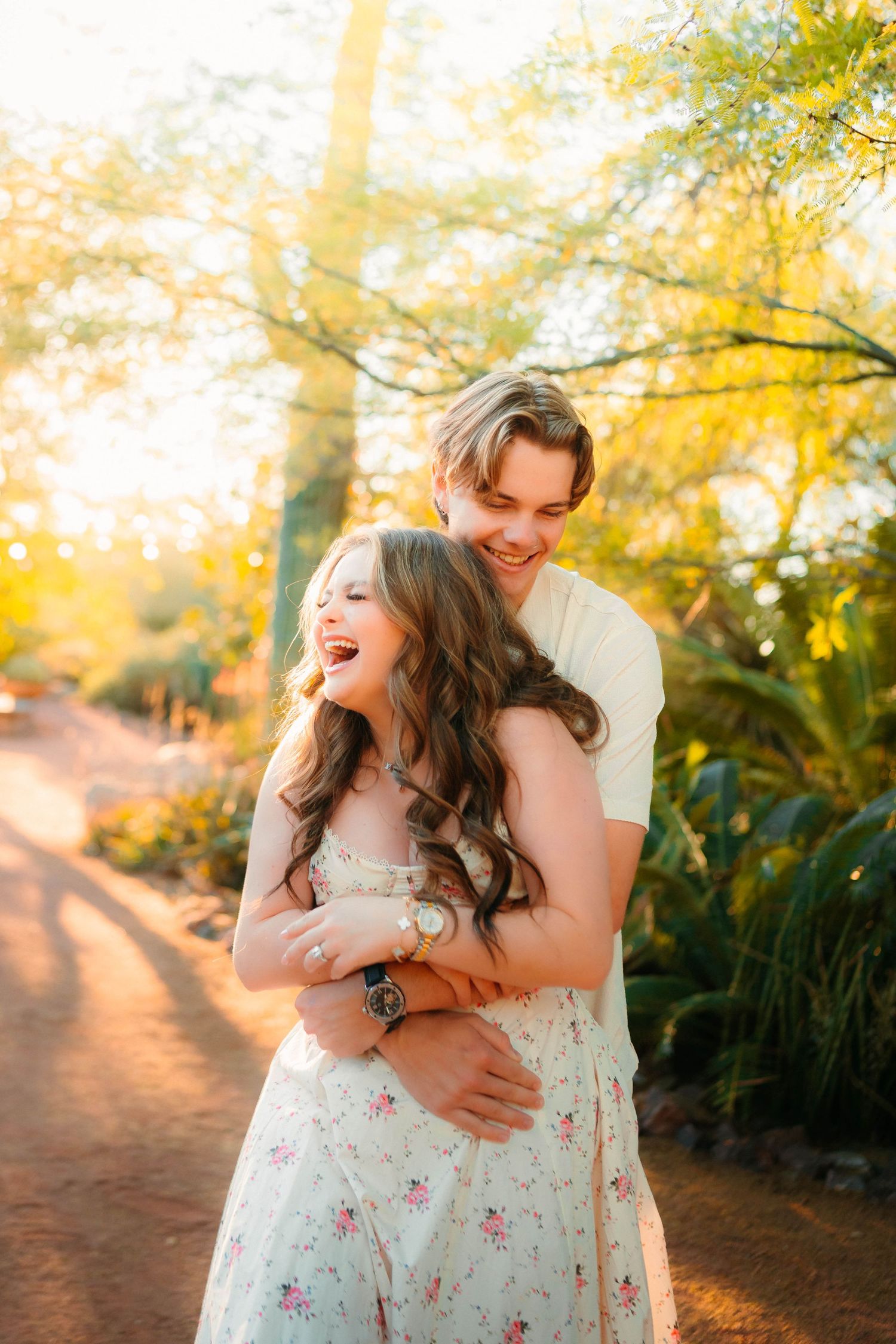 A joyful couple embraces outdoors in golden light, woman in floral dress laughing as man hugs her from behind.