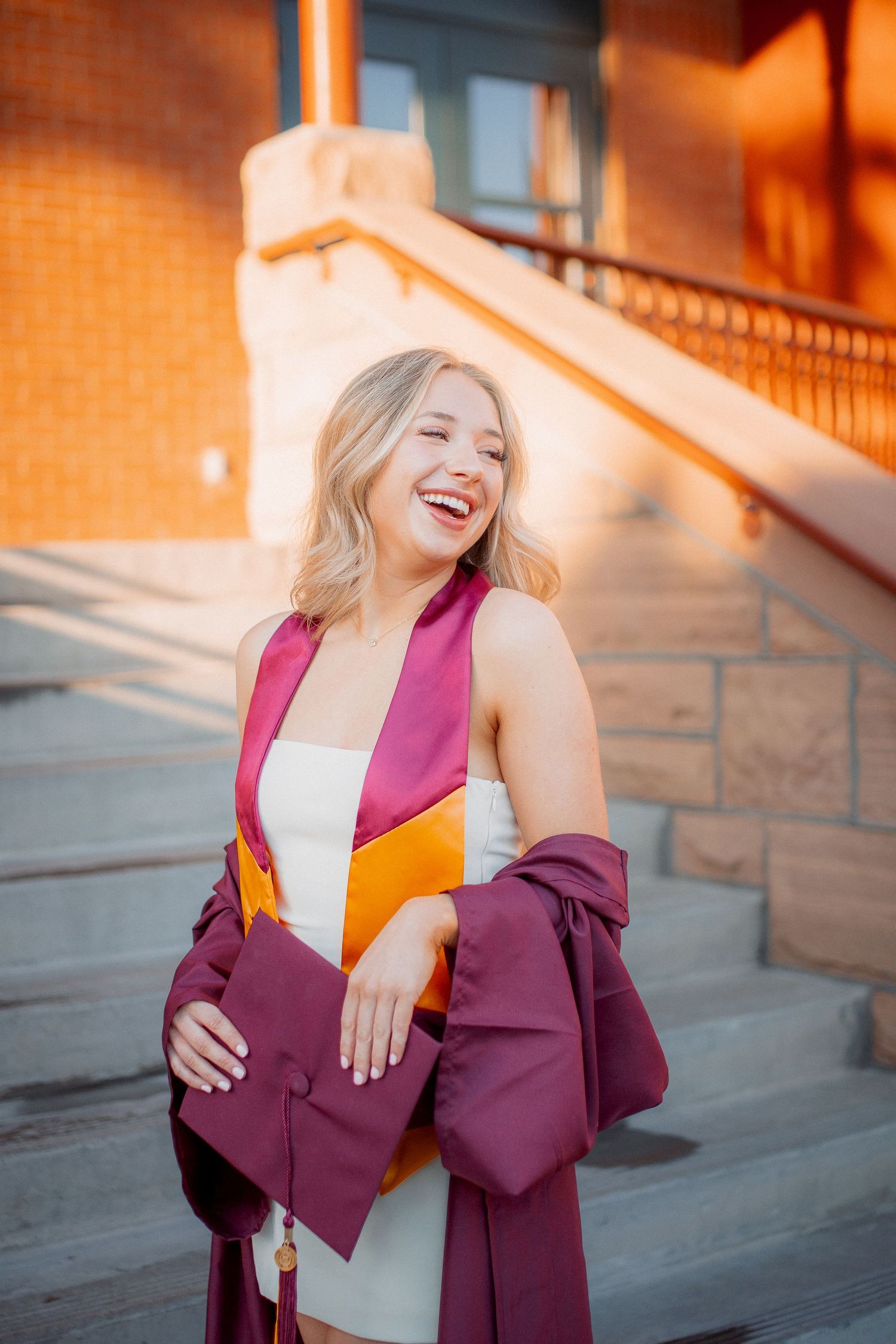 Smiling blonde graduate in maroon cap and gown poses on university steps in golden hour light.