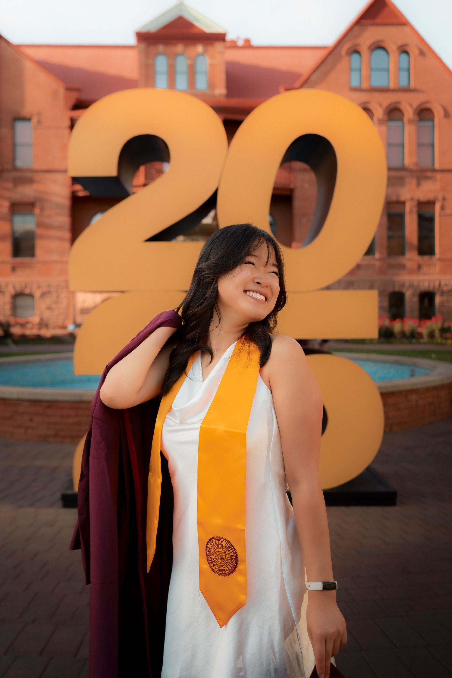 Happy graduate wearing white dress and gold stole posing in front of large 2025 sign on campus.
