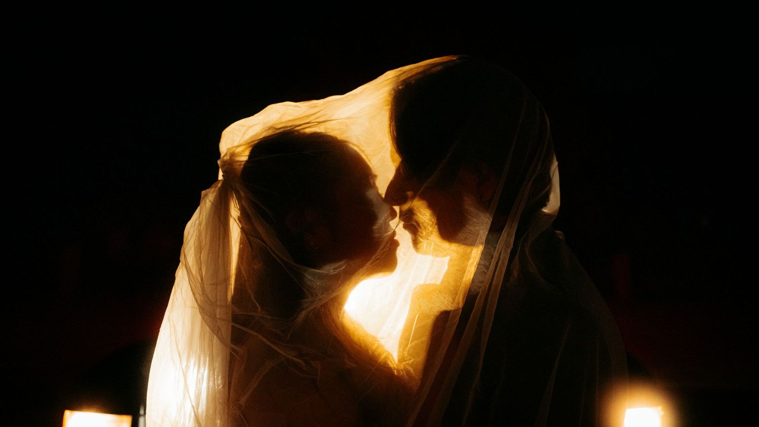 Bride and groom sharing an intimate moment under a veil in warm candlelight at night.
