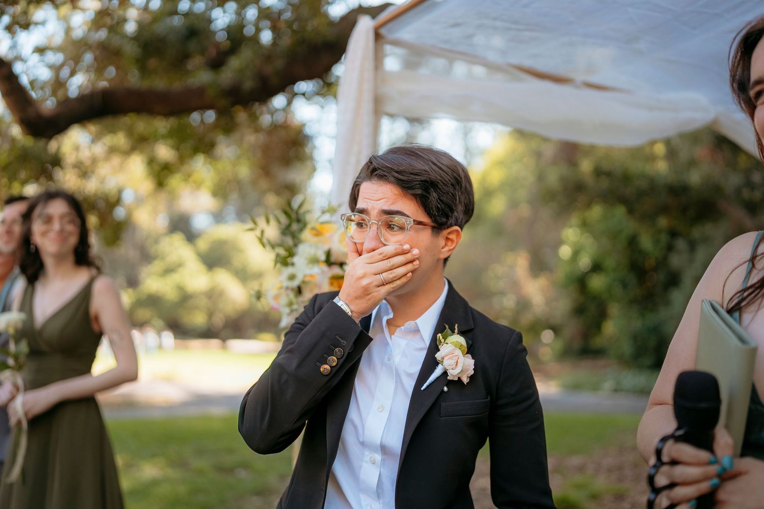 Emotional wedding guest covering their mouth in surprise during an outdoor garden ceremony.