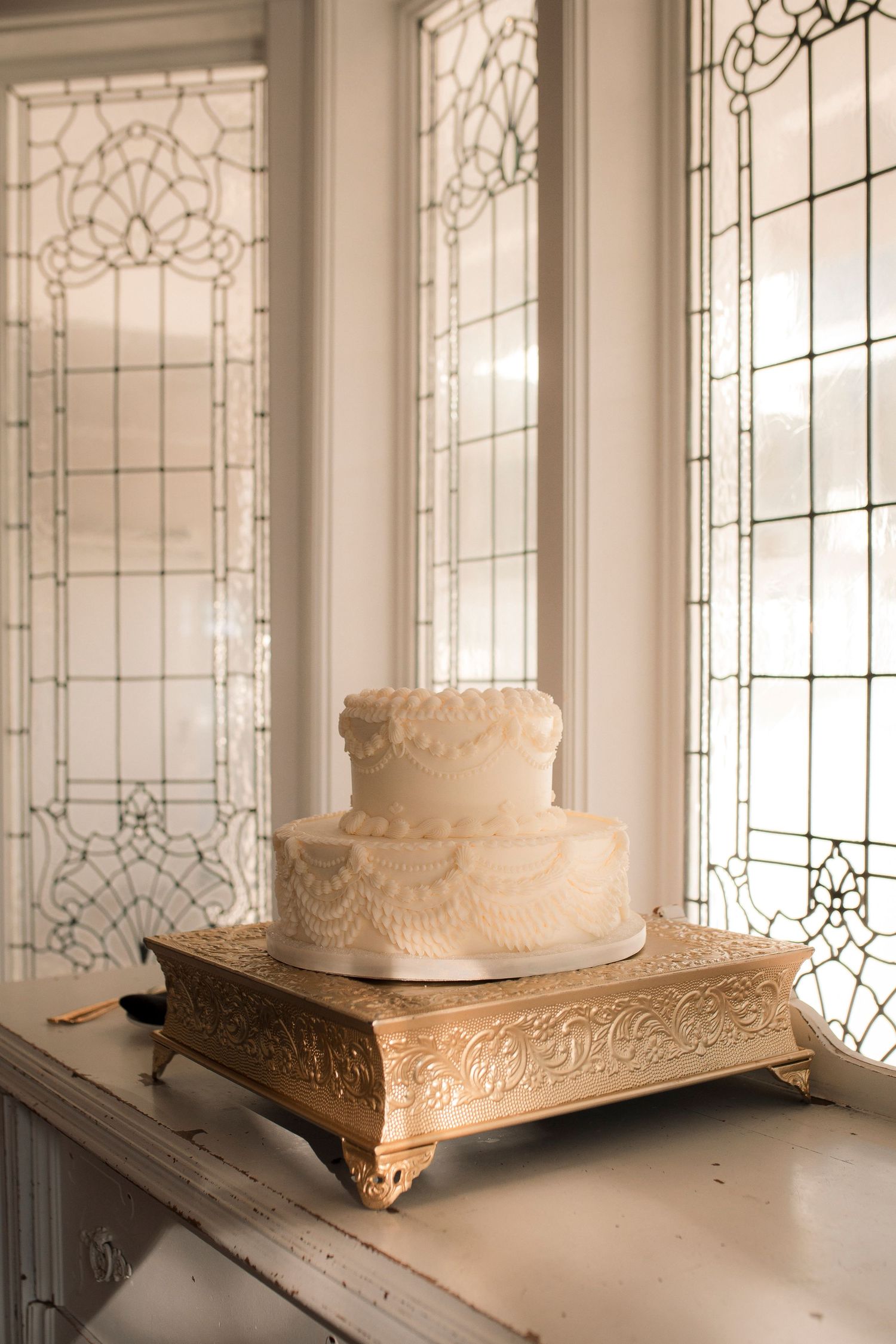 Elegant two-tier white lace wedding cake displayed on a gold ornate stand beside stained glass windows.