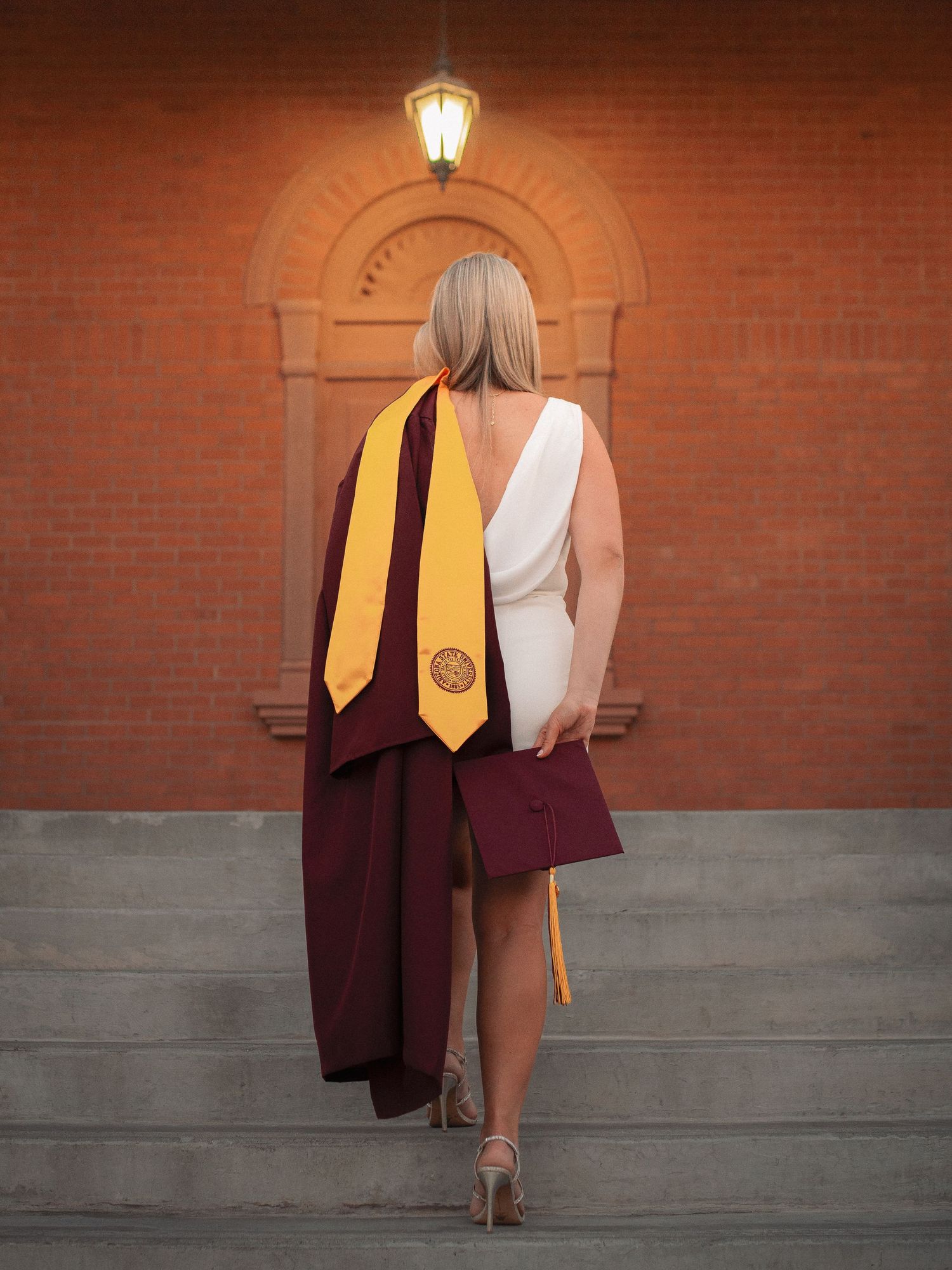 Graduate in maroon cap and gown with gold stole standing on steps before a brick archway.