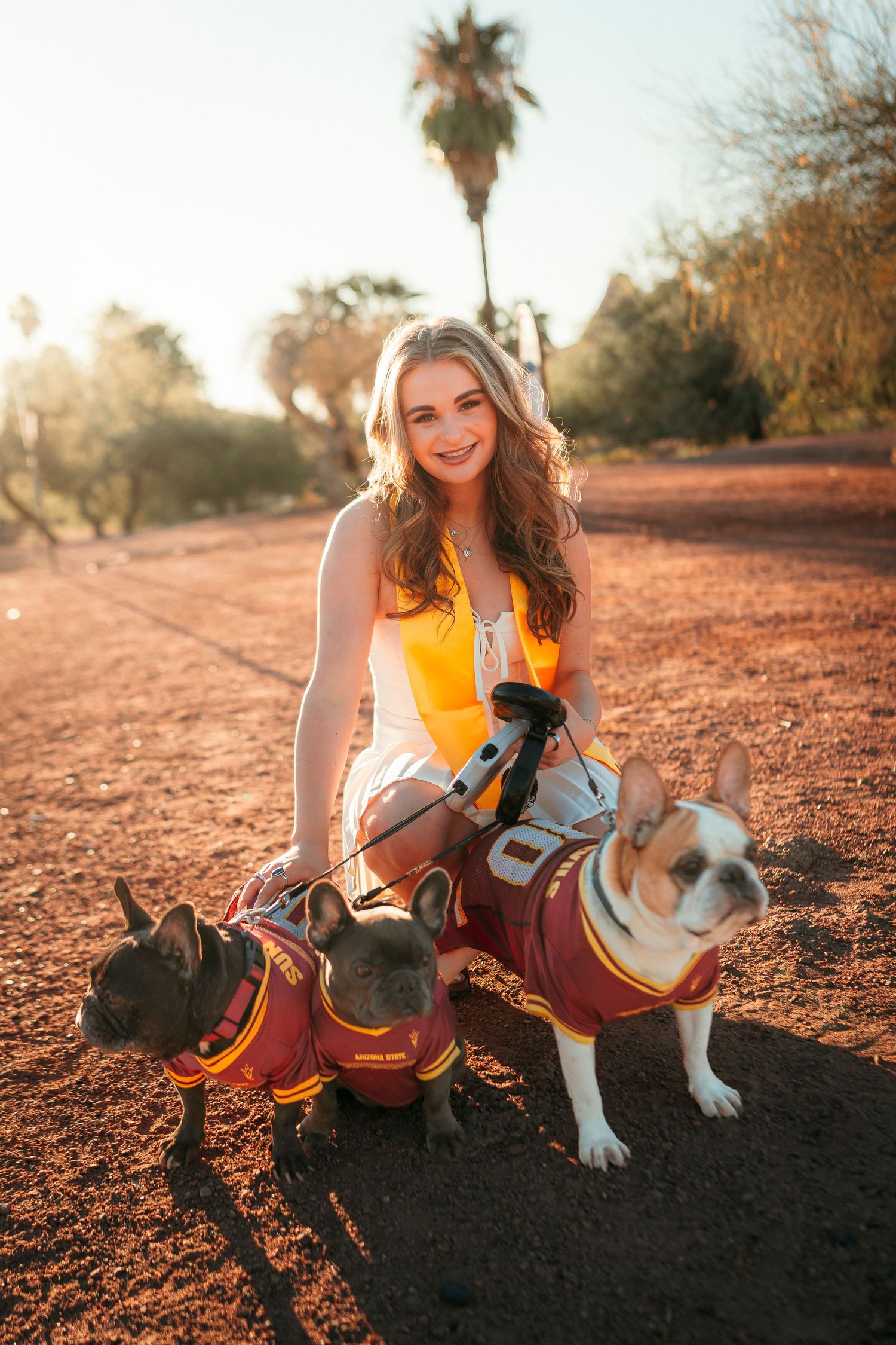 Smiling young woman posing with two French Bulldogs in ASU jerseys on red desert dirt path.
