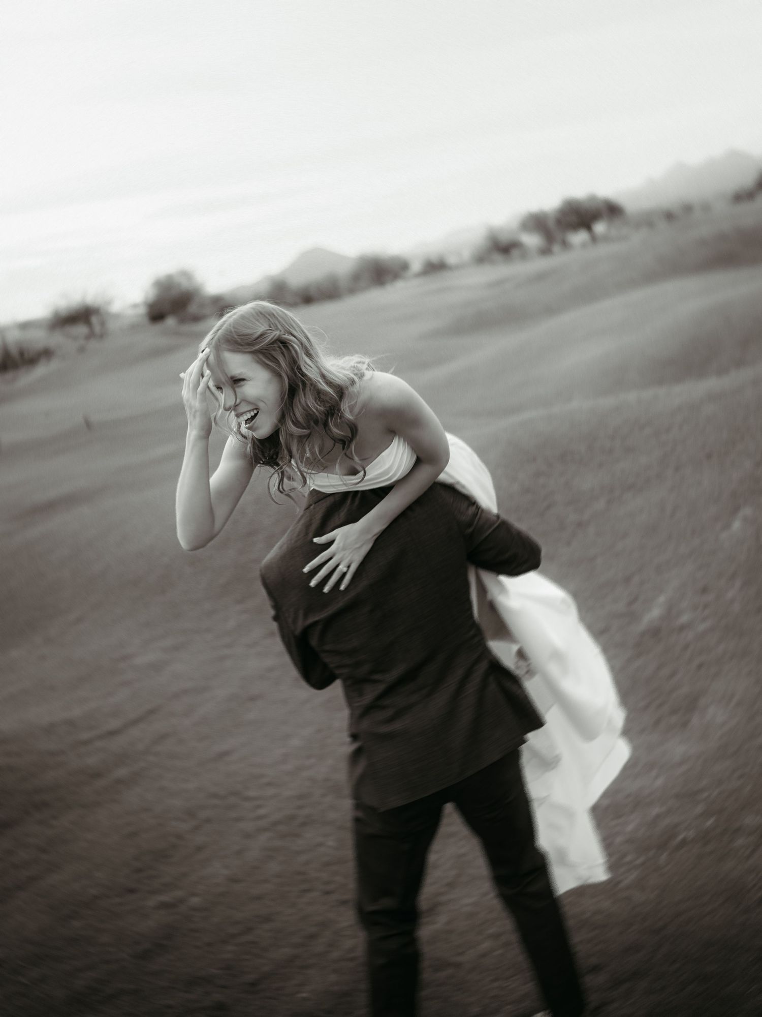 Groom carrying bride over his shoulder in a joyful black and white wedding photo on a scenic open field.
