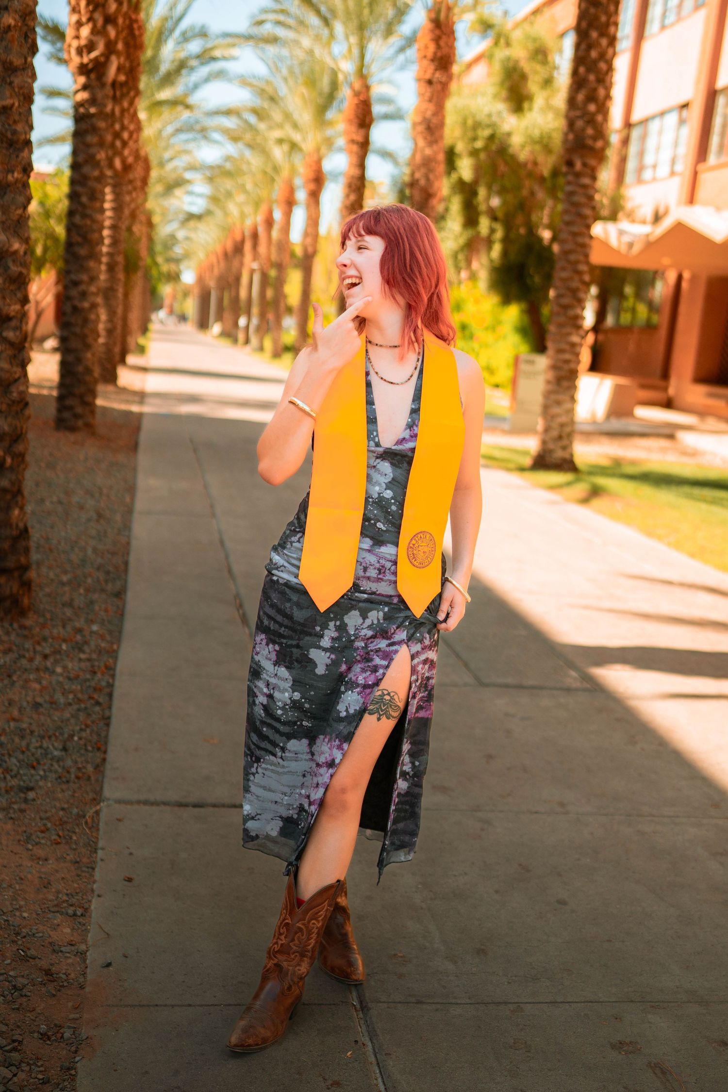 A smiling graduate in a floral dress and gold stole poses on a palm-lined campus walkway wearing cowboy boots.