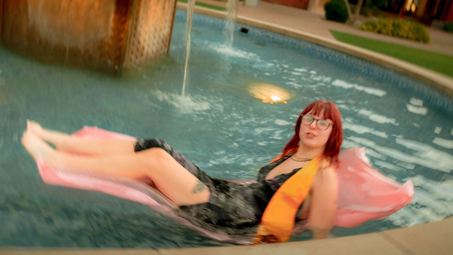 Young woman with red hair relaxing on pink float in outdoor pool at night.