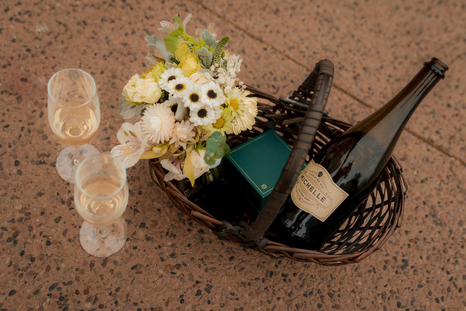Wicker basket with champagne bottle, flutes, and white floral bouquet on stone floor.