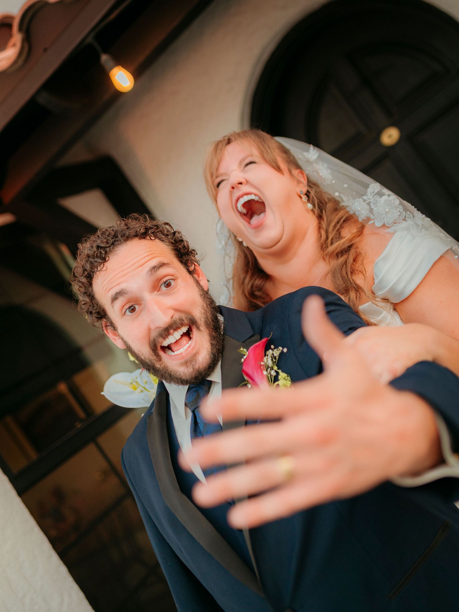 Joyful newlyweds celebrate at wedding, groom in navy suit holds bride in white dress as both laugh with excitement.