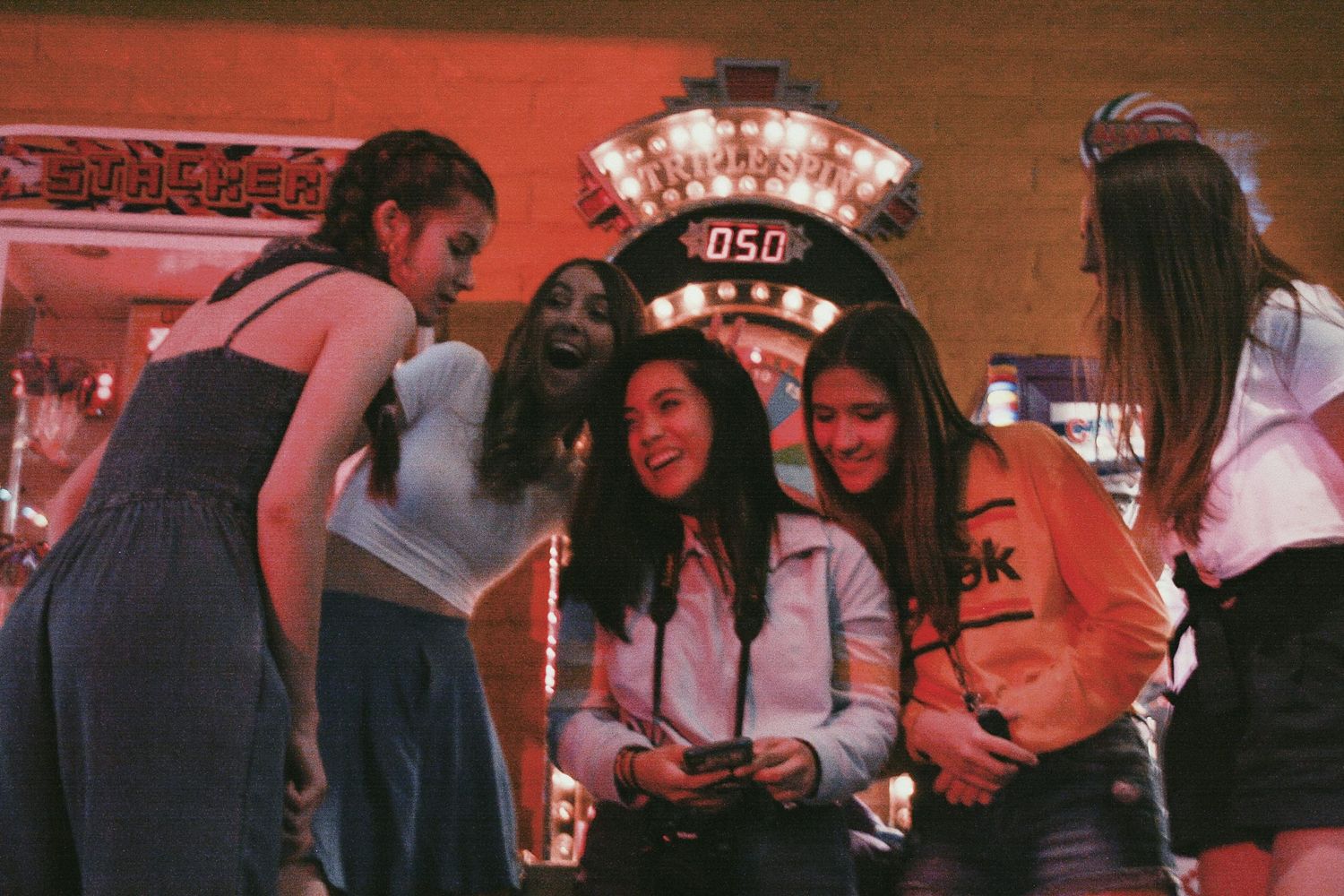 Group of young women laughing together near arcade game machine in colorful entertainment venue.