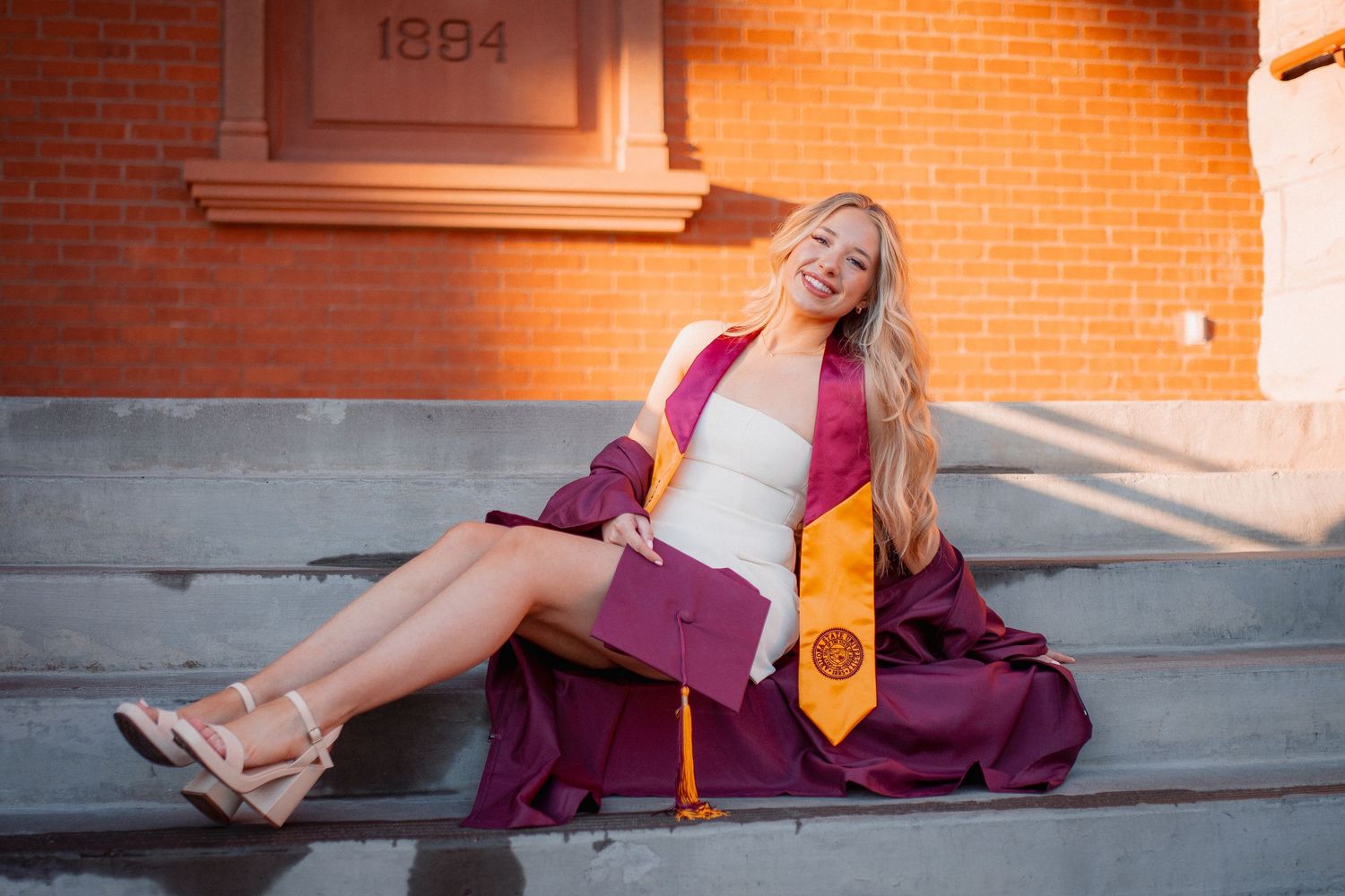 Smiling female graduate in maroon cap and gown sitting on steps with ASU colors.
