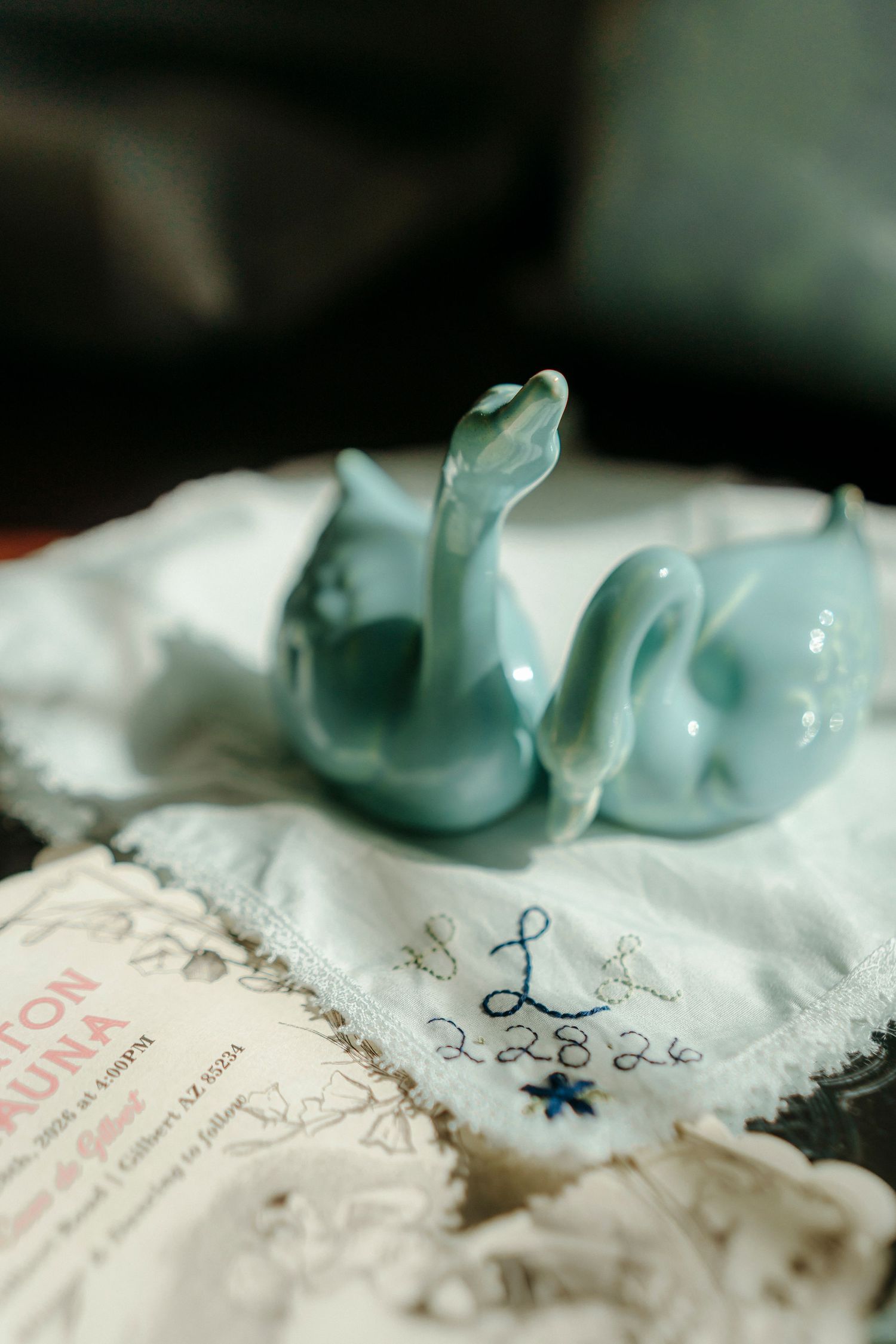 Blue ceramic swan figurines on embroidered handkerchief with monogram, beside wedding invitation card.