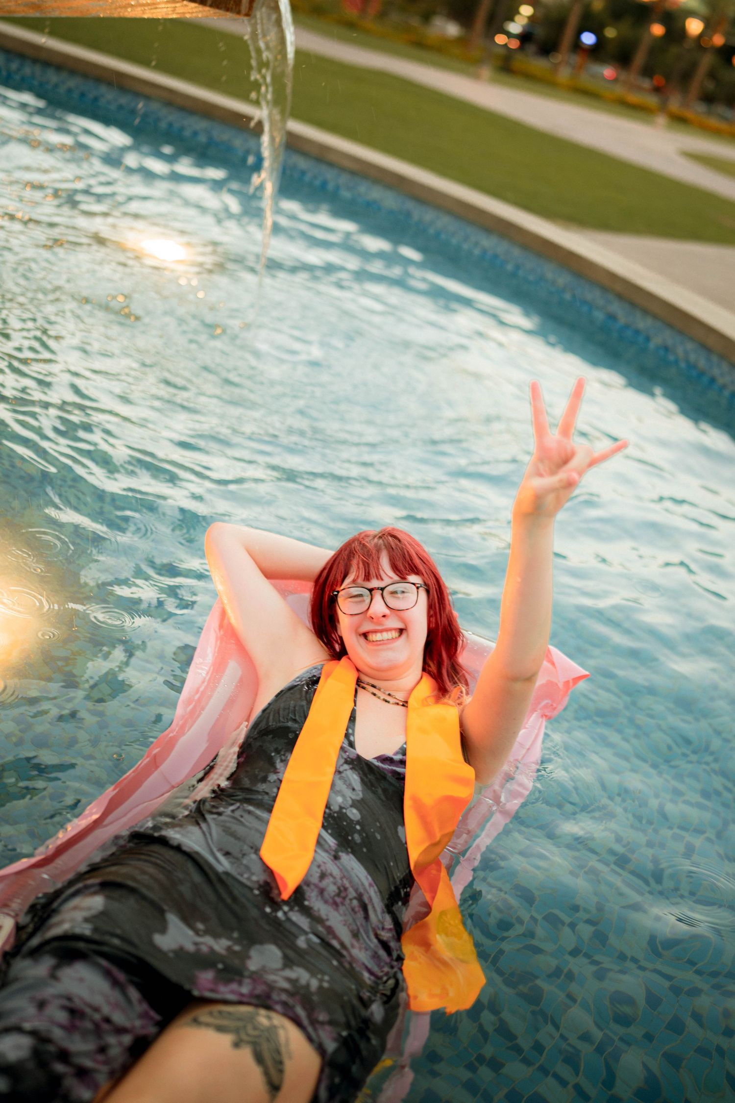 Happy redhead woman floating on pink raft in pool, flashing peace sign, wearing glasses and orange scarf.
